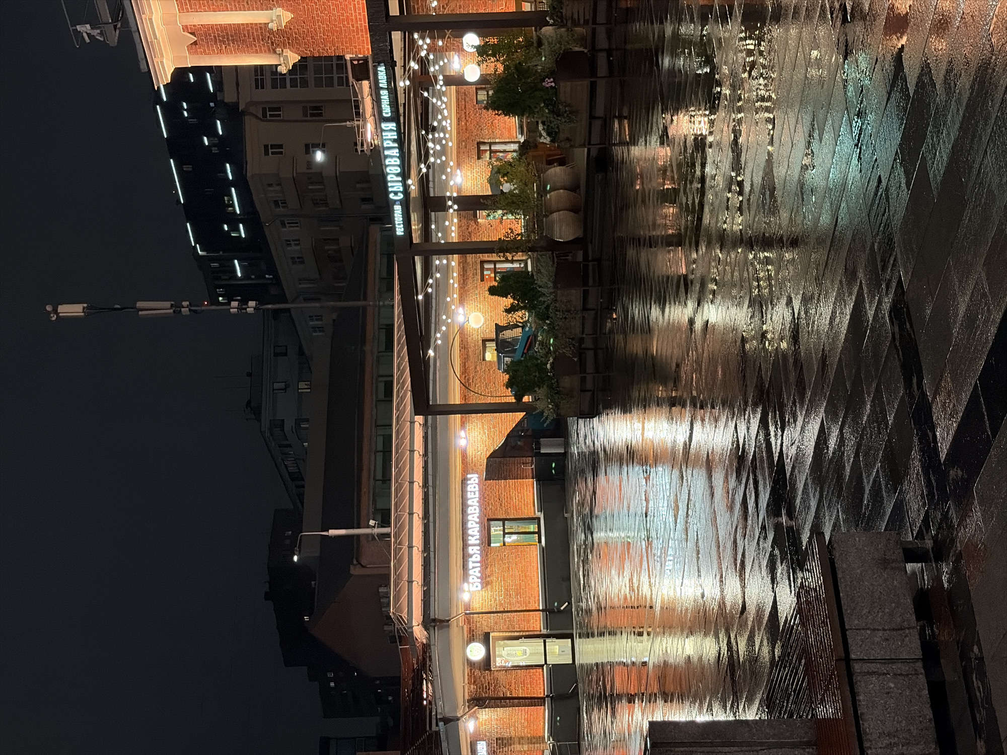 An outdoor view of a restaurant and its wet paved plaza at night. The restaurant, "BRATYA KARAVAEVY" (BROTHERS KARAVAEV), is constructed of red brick and has glowing signage. Numerous string lights are hung along the restaurant's canopy, illuminating the area. The plaza is covered in water, reflecting the lights and the surrounding buildings. In the background, several modern and older buildings are visible, silhouetted against the dark sky. A cell tower with multiple antennas stands tall to the left of the frame. The overall scene depicts a wet, urban environment at night, likely after a rain shower, creating a moody and atmospheric setting.