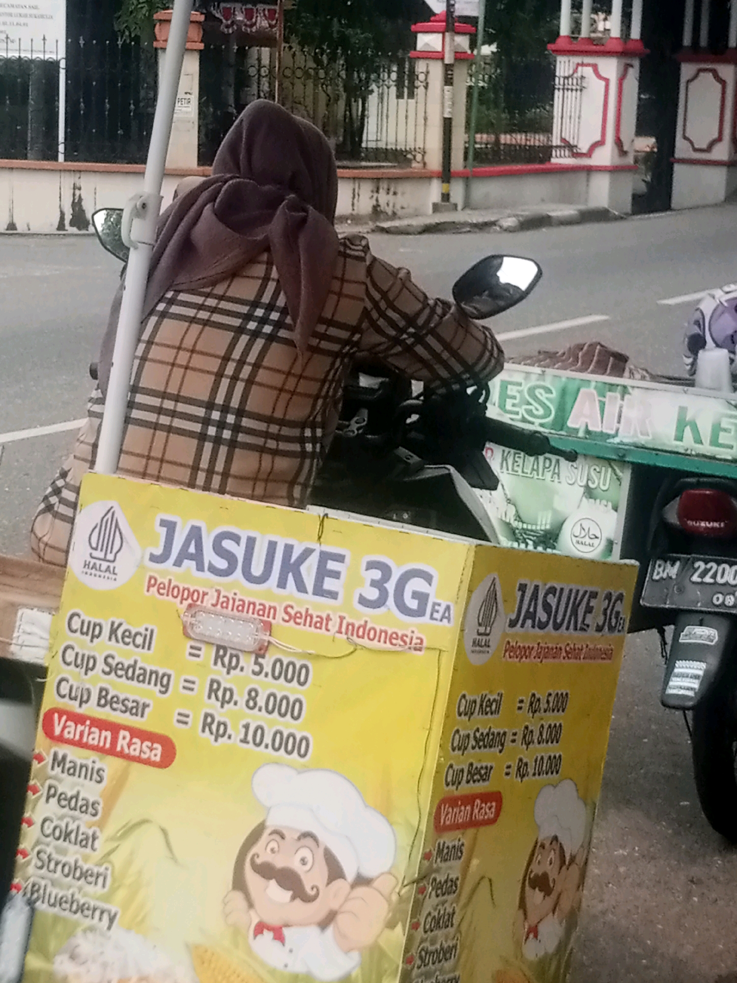 A person wearing a brown and black plaid jacket and a dark brown hijab is sitting on a motorcycle. In front of the motorcycle is a yellow sign with the text "JASUKE 3GEA" and "Pelopor Jajanan Sehat Indonesia". Below this are prices for different cup sizes: "Cup Kecil = Rp. 5.000", "Cup Sedang = Rp. 8.000", and "Cup Besar = Rp. 10.000". There are also listed flavors such as "Manis", "Pedas", "Coklat", "Stroberi", and "Blueberry". To the right of this sign is another identical sign. In the background, there is a green cart with the text "ES AIR KE" and "KELAPA SUSU". The environment appears to be a street in a city, with a sidewalk and a wall in the background. The license plate on the motorcycle reads "BM 2200 08". The context suggests this is a street vendor in Pekanbaru, Indonesia.