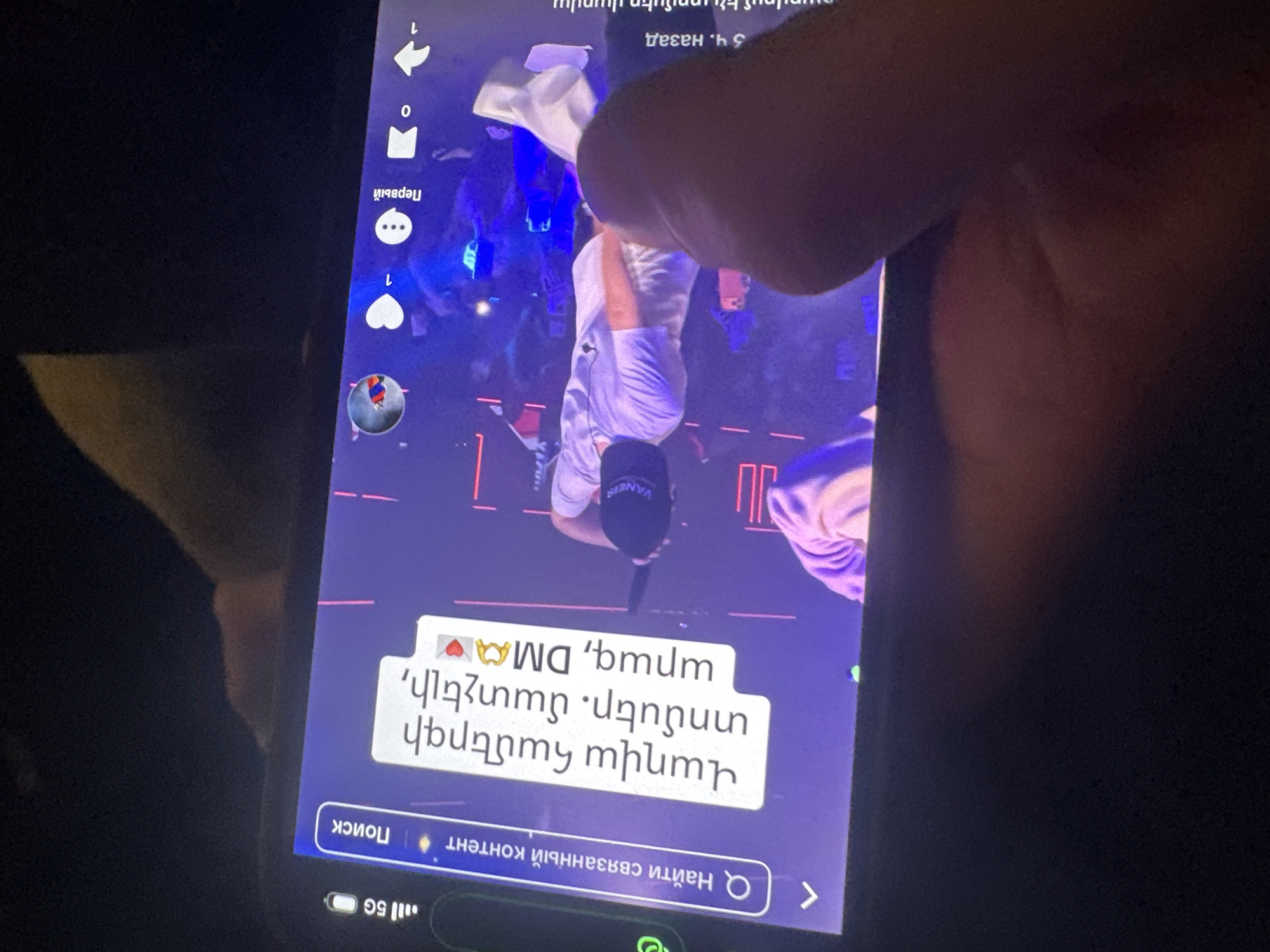 Here is a forensic description of the image:
The image shows a mobile phone screen displaying a vertical video. In the video, a person wearing a white outfit and a black cap is performing on stage in front of a crowd. The stage is illuminated with blue and red lights.
Overlaid on the video is a text box with Armenian text which roughly translates to "Tickets for tomorrow's concert. Affordable, fast, DMA". Below the video is a search bar with Russian text translating to "Find related content". The phone's interface shows icons for likes, comments, and sharing, along with network indicators showing a 5G connection. The phone is being held by a person, whose fingers and part of their hand are visible covering a portion of the screen. The rest of the environment is dark, suggesting the photo was taken in a dimly lit setting.