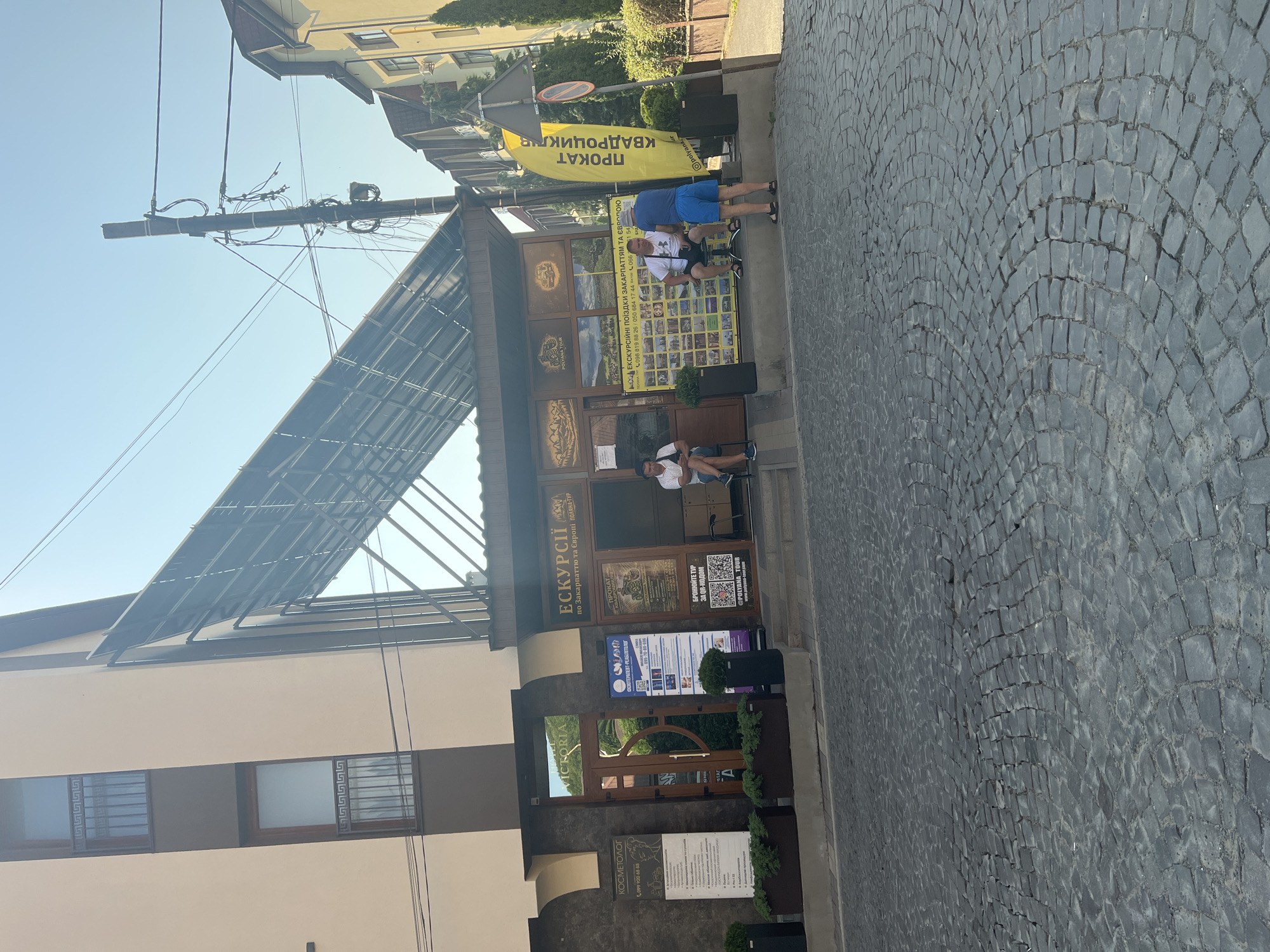The image shows the exterior of a building in the city of Сонячне, Ukraine. A cobblestone street is in the foreground, leading up to the entrance of a business. The building has a modern design with large windows and a slanted, glass-covered roof. Signs in Ukrainian advertise "ЕКСКУРСІЇ" (Excursions) and "ПРОКАТ КВАДРОЦИКЛІВ" (ATV rental). Three men are present: one is sitting on a bench to the left, another is sitting on a stool in front of the business, and the third is standing and talking to the man on the stool. The sky is clear and blue.