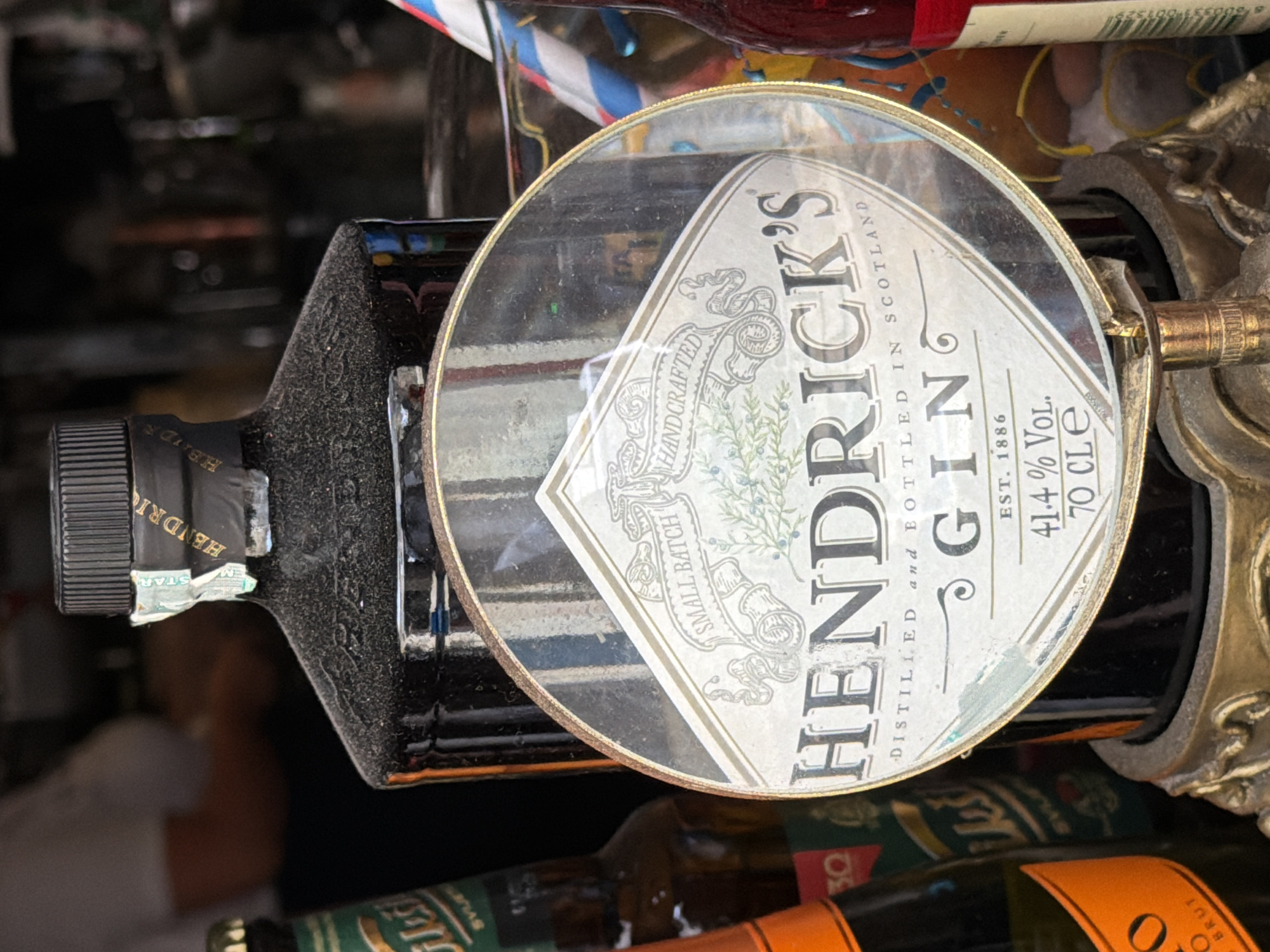 The image depicts a close-up view of a bottle of Hendrick's Gin, with its distinctive label prominently displayed and partially obscured by a magnifying glass. The bottle appears to be dark-colored and covered in a fine layer of dust, suggesting it may be on display in a bar or shop. The label reads "Hendrick's Gin," and further text indicates "Small Batch Handcrafted," "Distilled and Bottled in Scotland," "Est. 1886," "41.4% Vol.," and "70 Cle."
In the background, out of focus, are other bottles, one with a green label and text that appears to be in a Cyrillic script, and another with an orange label that is partially visible and reads "o BRUT." A striped straw is visible in a glass to the right of the gin bottle. The overall lighting suggests an indoor setting, possibly a dimly lit bar.
The provided location of Budva, Montenegro, is not visually discernible from the image itself. There are no visible people or distinct environmental features that confirm this location. The image focuses solely on the bottle and its label, with the surrounding elements serving as background context.