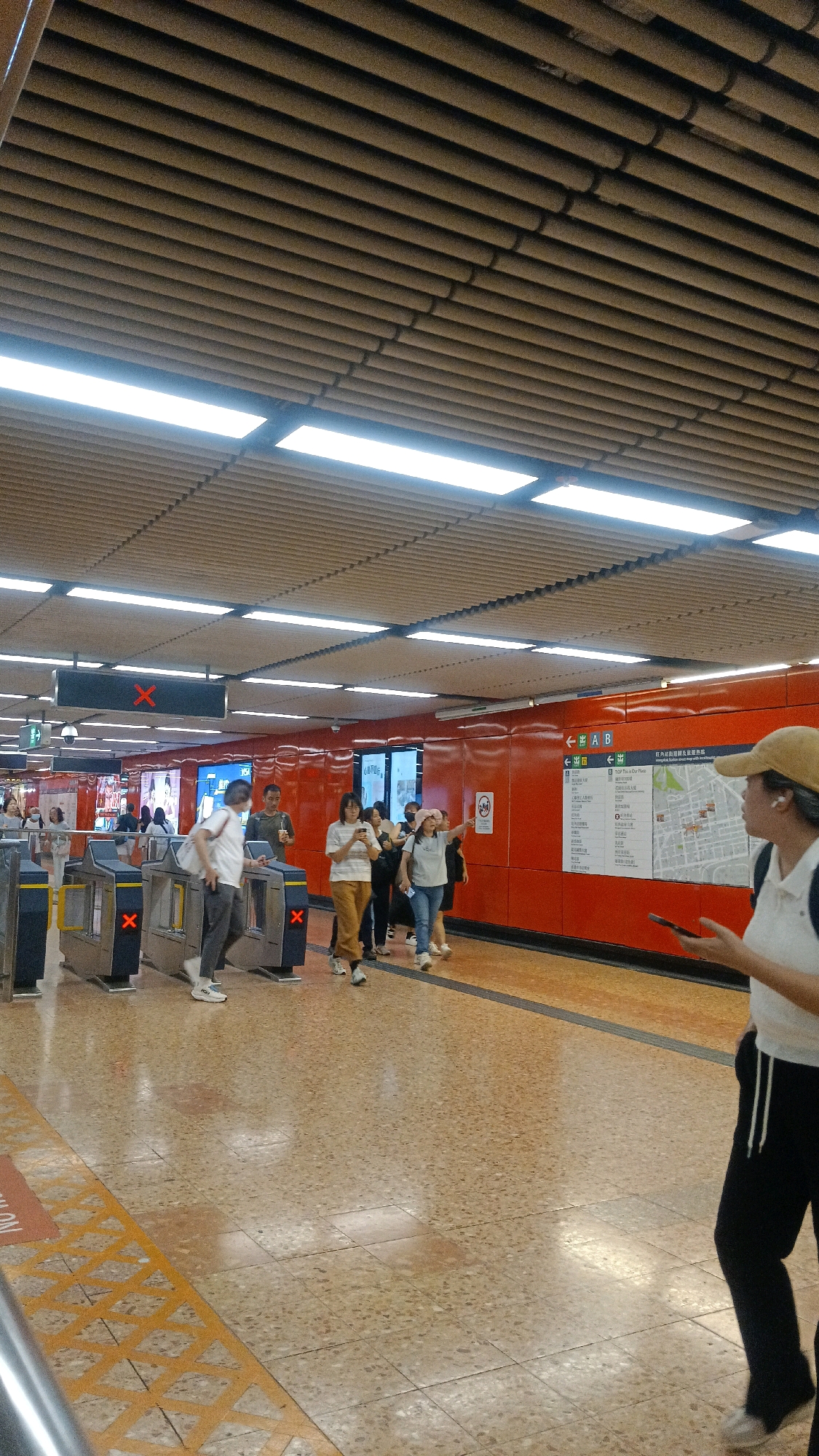 This is a medium shot of a busy subway station in Hong Kong, China. In the foreground, a person wearing a tan baseball cap and a white shirt is walking away from the camera, holding a smartphone. They are wearing black pants with white drawstrings. To their left, a group of people are walking towards a set of ticket gates. Some of these individuals are looking at their phones, while others are talking. The ticket gates are a dark gray and blue color, with red "X" symbols displayed on them.
The walls of the station are painted a bright orange, and a large map is mounted on the wall, indicating directions and locations within the city. The ceiling is made of horizontal wooden slats, with rows of bright fluorescent lights illuminating the space. The floor is tiled with a speckled beige and brown pattern. The overall environment suggests a public transportation hub in an urban setting.