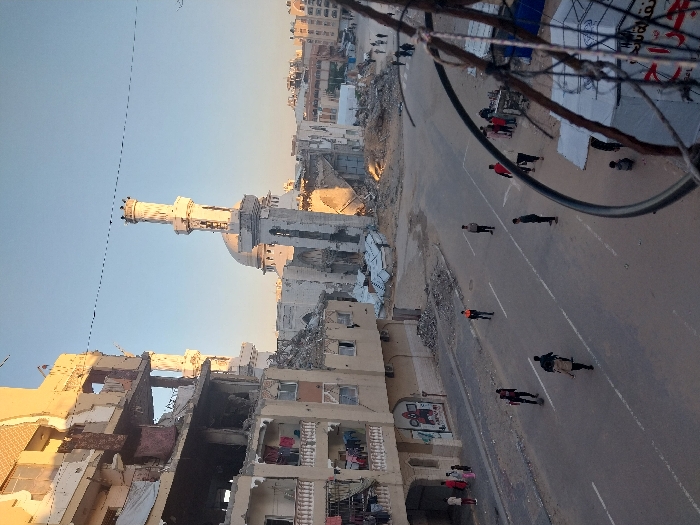 This image depicts a scene of extensive urban damage in Khan Yunis, Palestinian Territory. The central focus is a wide street flanked by heavily damaged buildings under a clear, bright sky, likely either morning or late afternoon given the lighting and shadows.
To the left, a multi-story residential building exhibits severe structural damage, with large sections of its facade and internal walls missing, exposing multiple rooms and balconies. Laundry is visible hanging from several damaged balconies. Piles of rubble are present at its base.
Dominating the mid-ground is a large, severely damaged mosque complex. Its prominent white dome is intact but surrounded by significant structural collapse, and a tall, slender minaret, also partially damaged, stands to its right. Extensive debris surrounds the base of the mosque.
The paved street running through the scene has clear white lane markings and is populated by numerous pedestrians. Groups of individuals, including men and children, are walking along the street and sidewalks. On the right side of the street, a group is gathered near what appears to be a small roadside stand or vendor. In the immediate foreground, to the bottom right, a metal frame with wires and some tangled material is partially visible, indicating the photo was taken from an elevated position. Below this, a section of a white makeshift tent structure can be seen. In the background, further damaged buildings and piles of rubble extend into the distance, with more pedestrians visible.