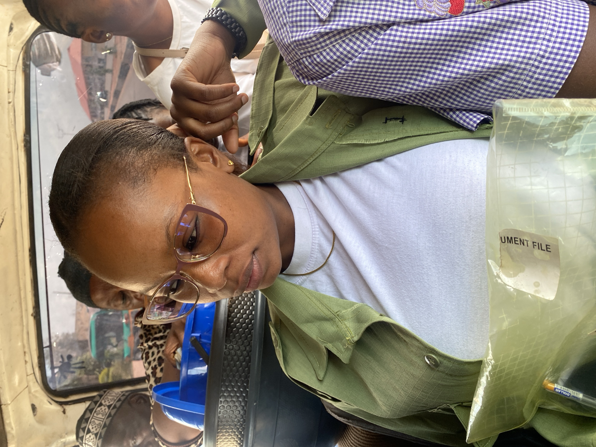 The image shows the interior of a vehicle in Gura Top, Nigeria. A woman with glasses and a white t-shirt under a light green shirt is looking down. A partially visible document file in a plastic bag with a pen inside is to her right. Two additional individuals are partially visible. One person is in a light purple checkered shirt, and another is in a white shirt.