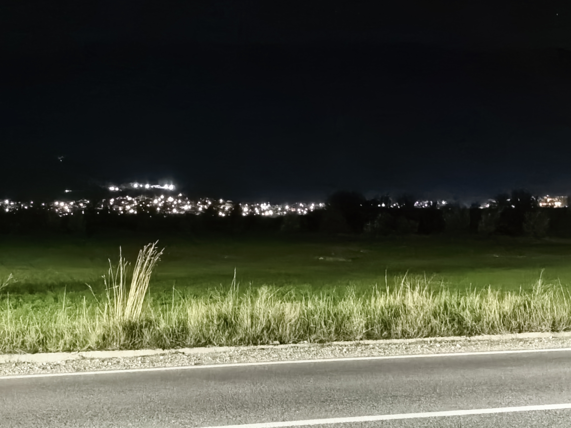 At night, a road cuts through the foreground, leading to a field of green grass. Tall, dry grass clumps stand out along the road's edge. In the distance, a town or city's lights are visible, scattered across the horizon. The sky above is dark. The location is Unknown, Unknown.