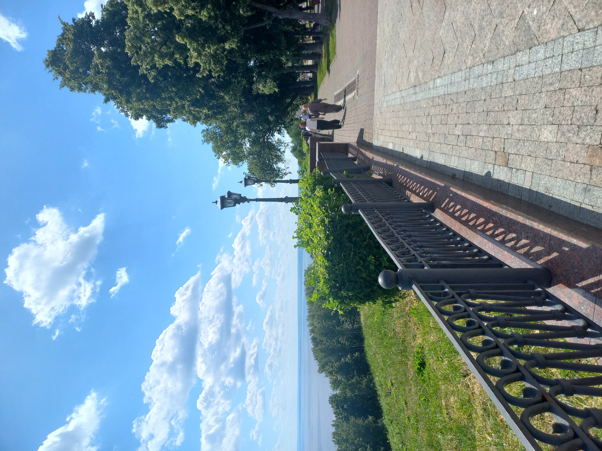 This is a photograph taken on a sunny day on the Volga River embankment in Ulyanovsk, Russia. A paved walkway lines the embankment, bordered by a decorative metal fence on the left. Beyond the fence, a grassy slope descends towards the wide expanse of the Volga River, with a forested shoreline visible in the distance.
On the walkway, two women are walking away from the camera, heading towards a line of large, mature trees that form a canopy over the path. Two old-fashioned lampposts are visible on the embankment, near a dense green bush. The sky is a bright blue, dotted with fluffy white clouds. The paving stones of the walkway are a mix of light grey and pinkish-brown, with a decorative border. The scene is well-lit, suggesting it is midday.