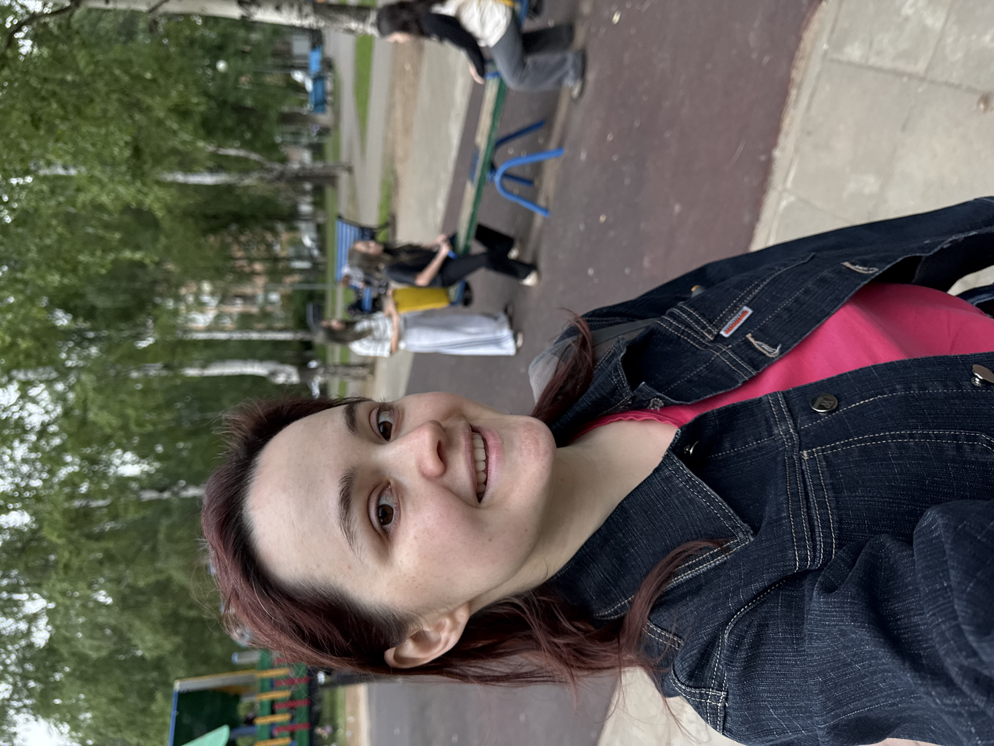 A selfie-style portrait captures a young adult female smiling directly at the camera within a park or public recreational area identified as being in Izhevsk, Russia.
**Main Subject:** The female subject, appearing to be in her 20s or early 30s, occupies the foreground. She has auburn hair, styled back from her forehead, with some strands loosely framing her face and resting on her shoulders. Her complexion is fair, displaying a scattering of light freckles across her nose and cheeks. Her brown eyes are open, and she exhibits a broad smile, revealing her upper teeth. She wears a dark indigo blue denim jacket with contrasting light-colored stitching details, a prominent collar, and silver-toned metal buttons on the front and a chest pocket. A vivid magenta t-shirt is visible beneath the jacket. A partially obscured red and white woven label bearing the text "WINDTONE" is affixed to the lower right side of her jacket.
**Background Environment:** The scene is an outdoor recreational area under diffused natural lighting, suggesting an overcast day. The ground directly behind the subject and extending into the midground is a dark reddish-brown, possibly asphalt or a rubberized playground surface. A grey concrete walkway runs along the bottom edge of the frame. The background is dominated by numerous tall deciduous trees with green foliage, many identifiable as birch trees due to their distinctive white bark. These trees form a dense canopy. In the far background, partially obscured by the trees, are multi-story residential buildings with light-colored facades.
**Other People and Objects:**
* In the midground, to the right of the subject, a blue-framed seesaw with a light brown wooden plank is in use.
* Two young females are seated on the seesaw: one in a dark long-sleeved top and dark pants on the left side, and another in a light-colored top and dark pants on the right side.
* A third young female, wearing a light-colored long-sleeved top (possibly striped) and a long, light-colored skirt or wide-leg pants, stands to the left of the seesaw, facing away from the camera.
* Several blue park benches are visible in the deeper background, with one obscured figure seated on a bench to the far right.
* On the far left side of the frame, a colorful playground structure with green, blue, and yellow elements is partially visible.