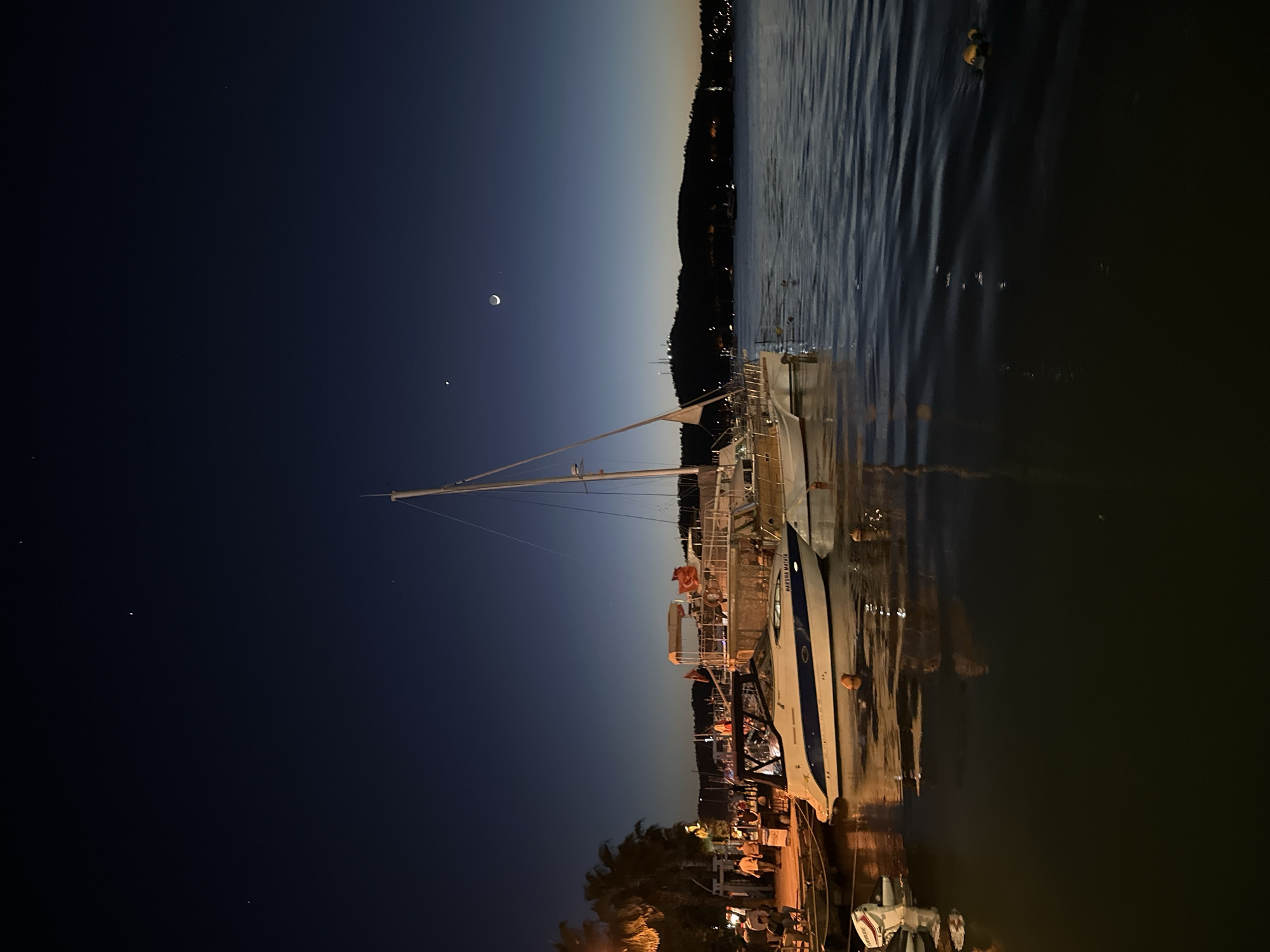 A nocturnal waterside scene in Fethiye, Turkey features boats moored near a lit harbor. The sky transitions from dark blue to light at the horizon with a crescent moon and visible stars. In the foreground, various boats and yachts are visible, illuminated by lights from the harbor. Reflections shimmer on the water's surface. A land mass with vegetation and buildings is silhouetted in the distance. The city lights create a vibrant atmosphere, contrasting with the dark night sky.