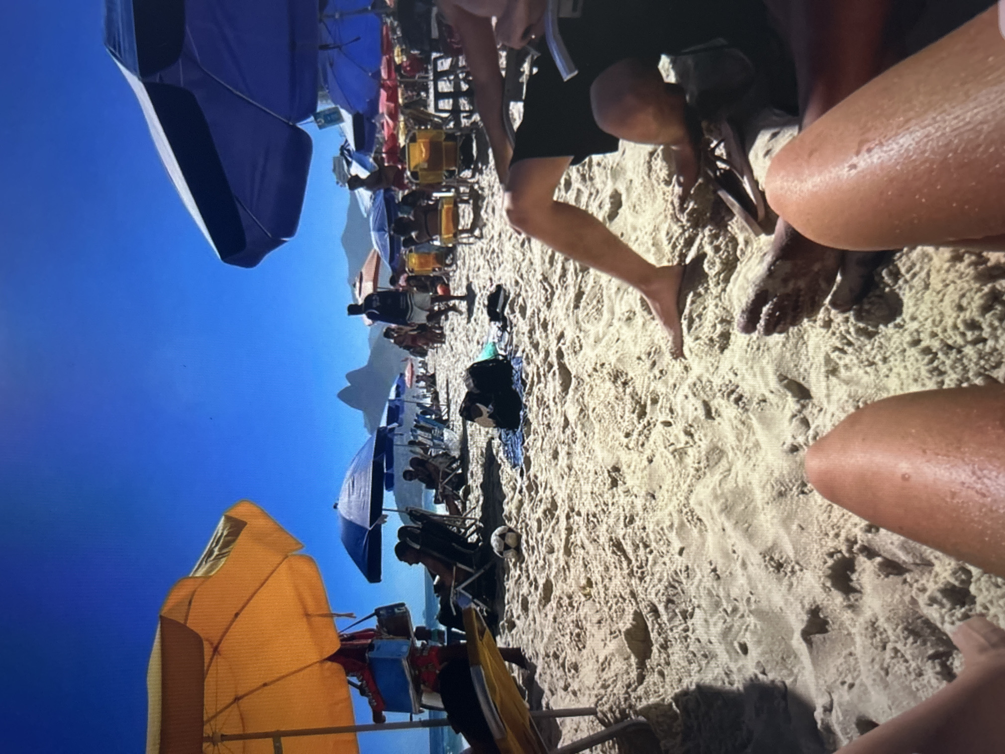 In an outdoor, daytime scene on a sandy beach under a clear blue sky, multiple beach umbrellas, predominantly orange and blue, provide shade. Several people are visible, some seated in beach chairs, others standing or walking along the shore. A soccer ball rests on the sand in the mid-ground. In the background, mountains are visible. The foreground is occupied by the legs and feet of two individuals. The location is specified as Unknown, Unknown.