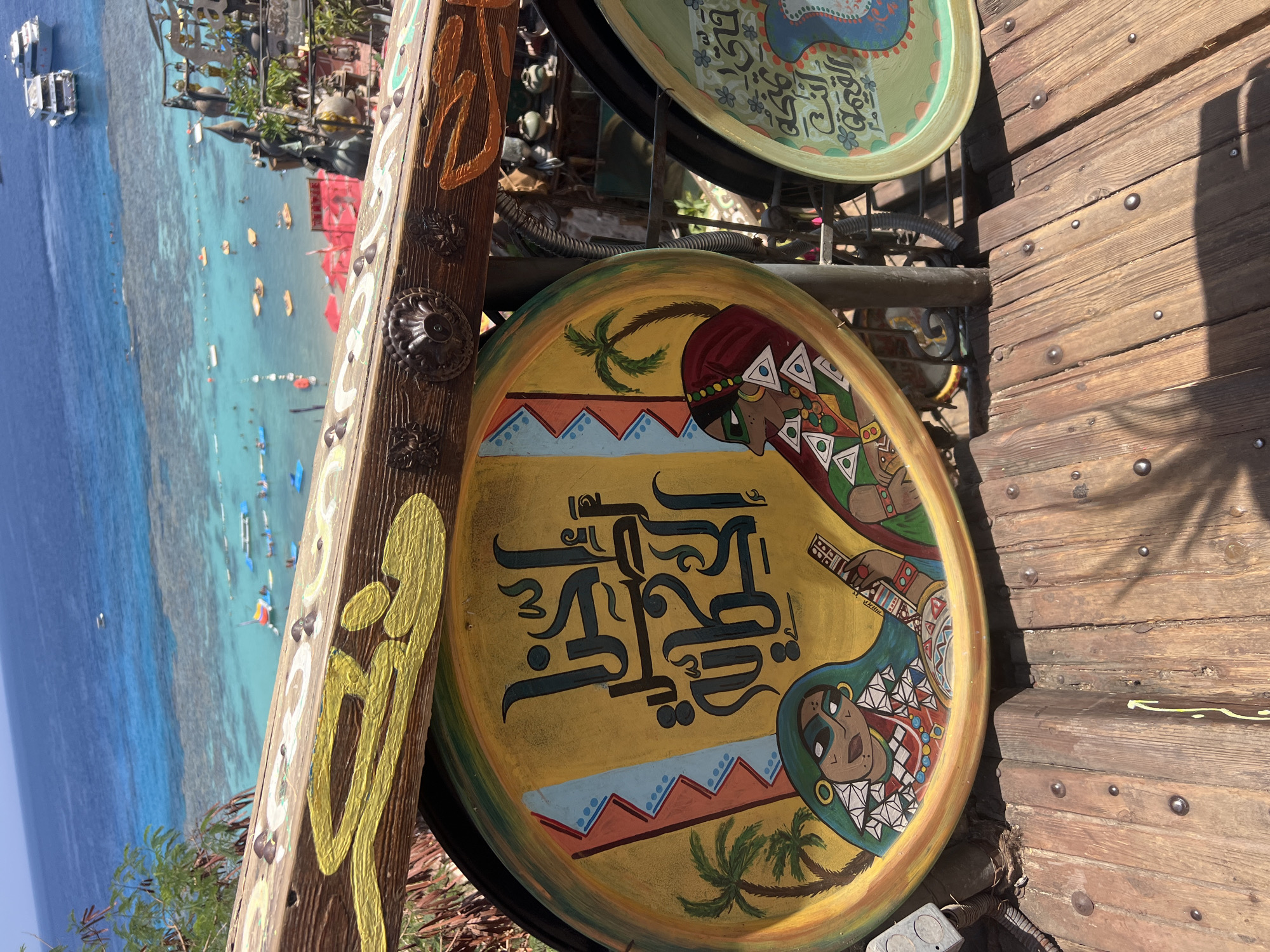 The image displays an outdoor scene overlooking a bright blue ocean with several boats visible. In the foreground, there are two large, round decorative plates with colorful Arabic calligraphy and painted figures. The plate on the left is yellow and features two women in traditional attire, one playing a stringed instrument. Palm trees and geometric patterns are also depicted. The plate on the right is green and shows more Arabic script and decorative elements. These plates are mounted on a wooden structure that appears to be part of a railing or balcony, with visible metal accents and rivets on the wooden surface. The backdrop is a clear blue sky and the turquoise waters of the sea, with a coral reef visible beneath the surface. The location is identified as Sharm El Sheikh, Egypt, suggesting a scenic tourist spot.