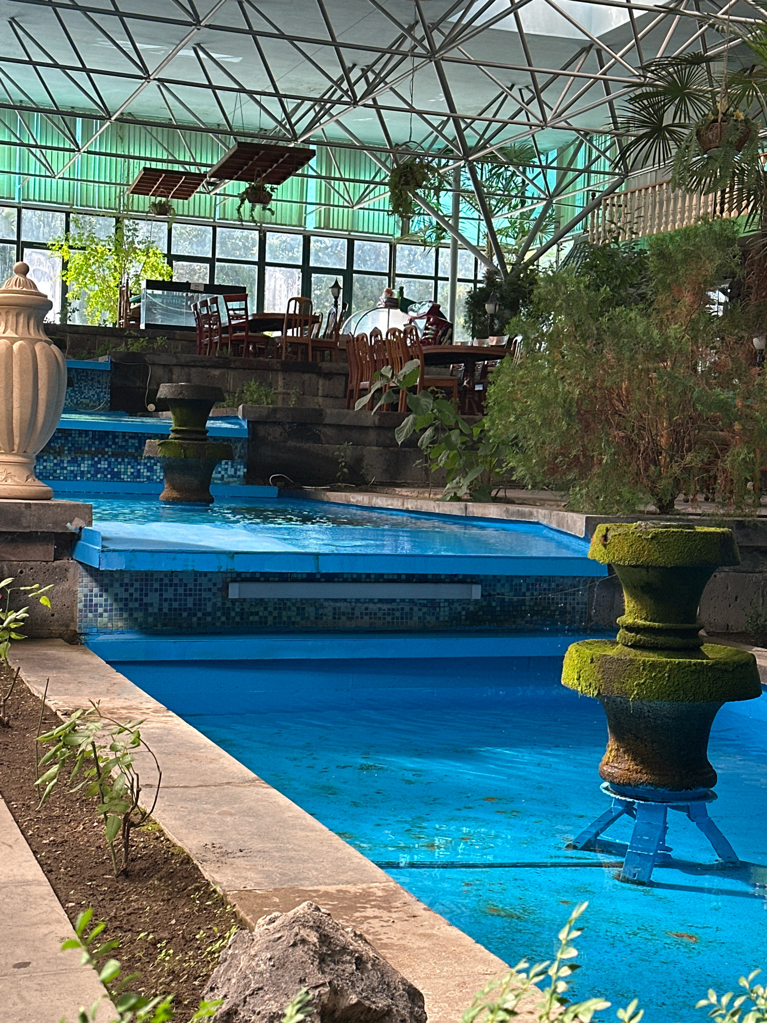 The image shows an indoor courtyard with a tiered blue pool and multiple fountains. The pool area is surrounded by stone walls and planters with small green plants. In the background, there are several dining tables and chairs arranged under a large, geodesic dome-like glass roof structure. Lush greenery, including trees and hanging plants, is visible throughout the space, creating a conservatory-like atmosphere. The overall setting appears to be a restaurant or cafe with an indoor garden and water features. The location is identified as the "New Gyumri Hotel" in Gyumri, Armenia. There are no people clearly visible in the frame.