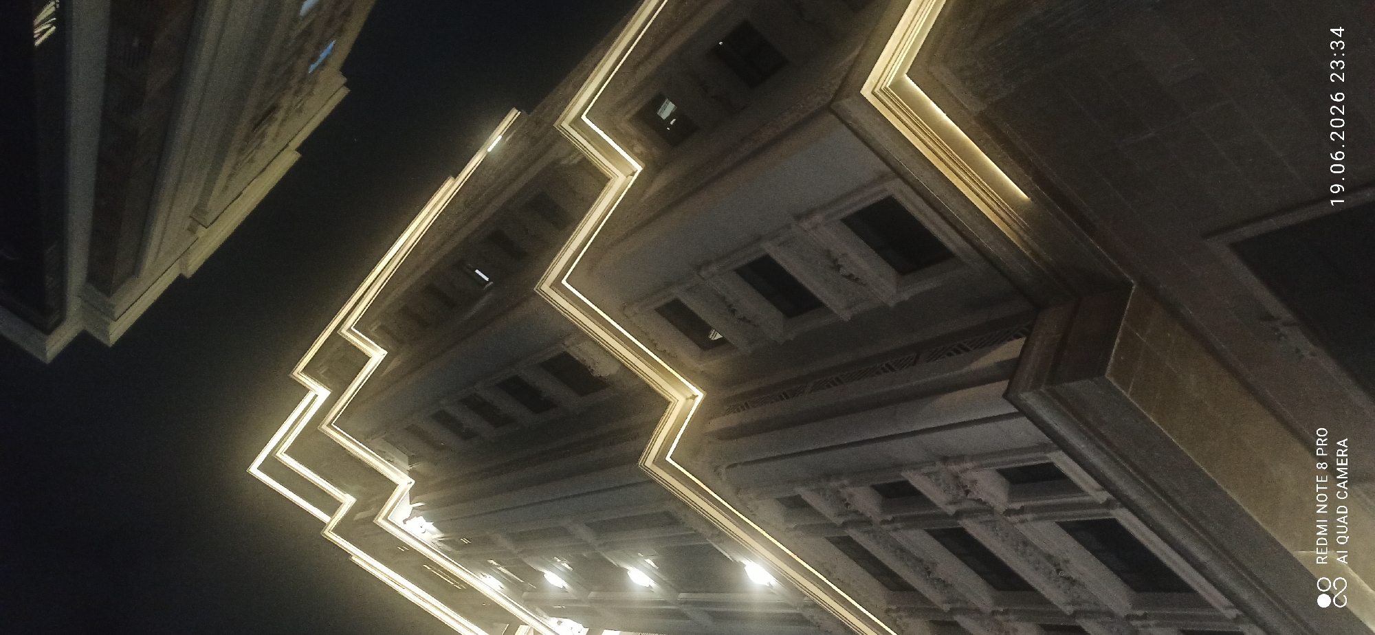 An upward-angle shot shows the exterior of a modern building at night. The building is illuminated by warm, yellow accent lighting that follows the sharp, angular lines of its facade, creating a striking geometric pattern against the dark sky. The architecture features multiple levels with rows of dark windows, some of which show internal lighting. Ornate decorative elements are visible on some of the building's ledges and window frames. In the upper right quadrant of the image, another building facade, also illuminated, is partially visible, suggesting an urban setting. The bottom left corner of the image contains a watermark displaying "REDMI NOTE 8 PRO" and "AI QUAD CAMERA," and the bottom right corner shows a timestamp: "19.06.2026 23:34."