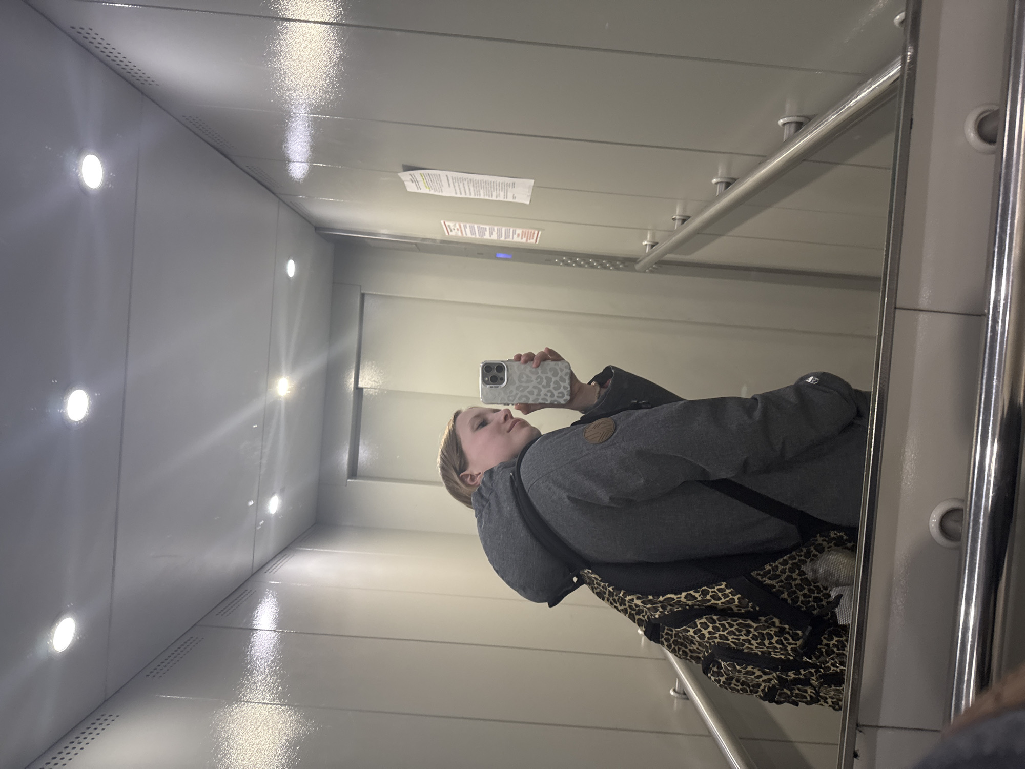 The image shows a selfie taken by a person reflected in the highly reflective interior of an elevator. The setting is an elevator, likely in Chaikovsky, Russia, given the context.
A person, appearing to be female, is visible in the reflection, holding a white smartphone with a distinctive leopard print case to capture the image. She is wearing a grey winter jacket with a brown circular patch on the left sleeve and a black strap from a bag, which also has a leopard print pattern, visible across her chest and back. Her expression is neutral, focused on the phone screen.
The elevator interior is characterized by shiny, light grey or metallic panels on the walls and ceiling, which act as reflective surfaces, creating a mirrored effect and reflecting the bright, recessed circular lights embedded in both the ceiling and walls. Small, perforated vent holes are visible along the top edges of the walls and where the walls meet the floor. Polished silver handrails extend horizontally along the walls. Above the person's reflection, two paper notices are affixed to the elevator wall; one is larger and white with yellow highlighted lines, and the other is smaller with red headings. The text on these notices is too indistinct to read, but they appear to be informational or safety instructions. A small, blue digital display is also visible above these notices. The floor also appears to be a reflective material, mirroring the overhead lights. The overall impression is a modern, well-lit, and clean elevator.