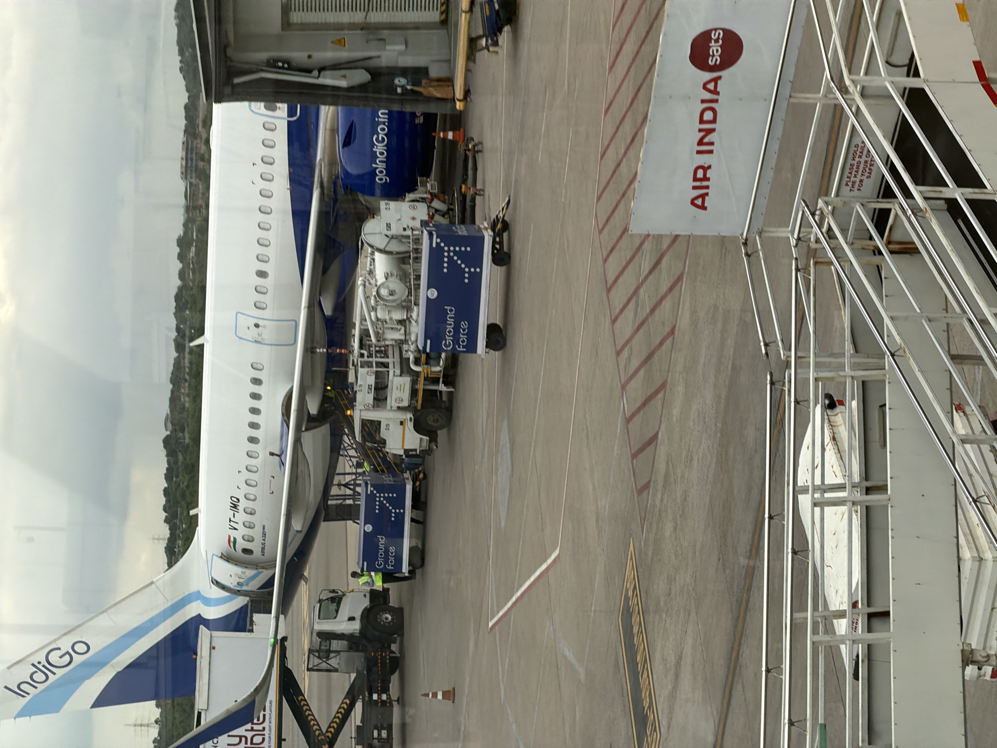 The image captures an outdoor scene at an airport in Mamidipally, India, likely during daylight hours under an overcast sky. In the foreground, a white airstair with metal railings leads upwards. A sign displaying "AIR INDIA sats" is mounted on the structure. Below the airstair, a concrete tarmac is visible with red and white markings. Several vehicles are present on the tarmac. A white truck with a loading ramp is positioned to the left. Adjacent to it is a blue baggage cart with the words "Ground Force" and an arrow design. Further to the right, another white utility vehicle is parked. In the background, a large airplane with "IndiGo" branding on its tail is parked at a gate. The fuselage of the plane is white with a blue stripe. Part of a jet bridge or gate structure is visible on the right side of the image. The overall environment suggests an operational airport setting.