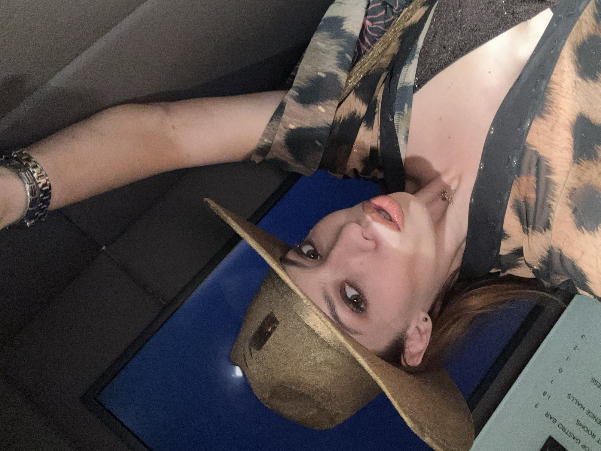 The image is a selfie taken by a woman wearing a golden-brown fedora hat and a leopard-print top. Her left arm is raised above her head, and she is wearing a silver watch on her left wrist. The background appears to be a dark, possibly upholstered surface, with a blue pane of glass or window to her left. The woman is looking towards the camera with her mouth slightly open, and she is wearing makeup, including lipstick. A portion of a light blue sign with text is visible in the lower left corner of the image, containing words such as "TOP GASTRO BAR", "T ROOMS", "DENCE HALLS", and numbers such as "9", "1-8", "1", "0", "2", "-2", "-3". The text and the presence of a menu or information board suggest that this image was taken in a public establishment, potentially a hotel, restaurant, or bar. Without further information, it is impossible to determine the specific location in Sofia, Bulgaria.