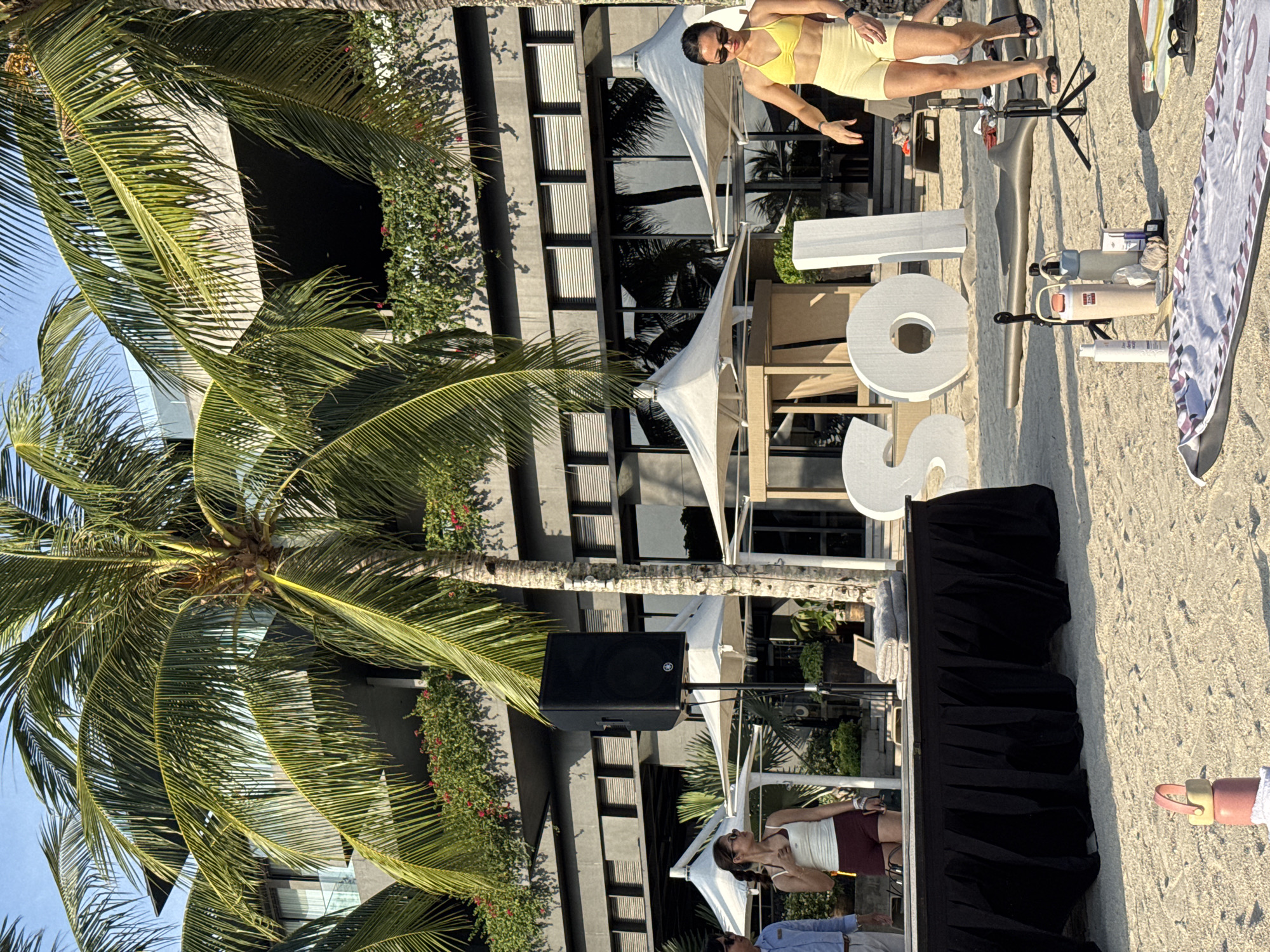 The image depicts an outdoor scene on a sandy beach in Samal, Philippines, likely a resort or event space. In the foreground, a striped yoga mat lies on the sand, with several water bottles and a tripod. Large white letters spelling "SOL" stand in the sand behind the mat. A woman in a yellow bikini and shorts is walking towards the right side of the frame, and another woman in a white tank top and maroon shorts is standing near a black table draped with a black cloth. A large black speaker on a stand is positioned to the left of the table. Behind the subjects, a modern building with a grey facade and large windows is visible, flanked by palm trees and lush greenery. White umbrellas are set up near the building. The lighting suggests it is daytime, with the sun casting shadows.