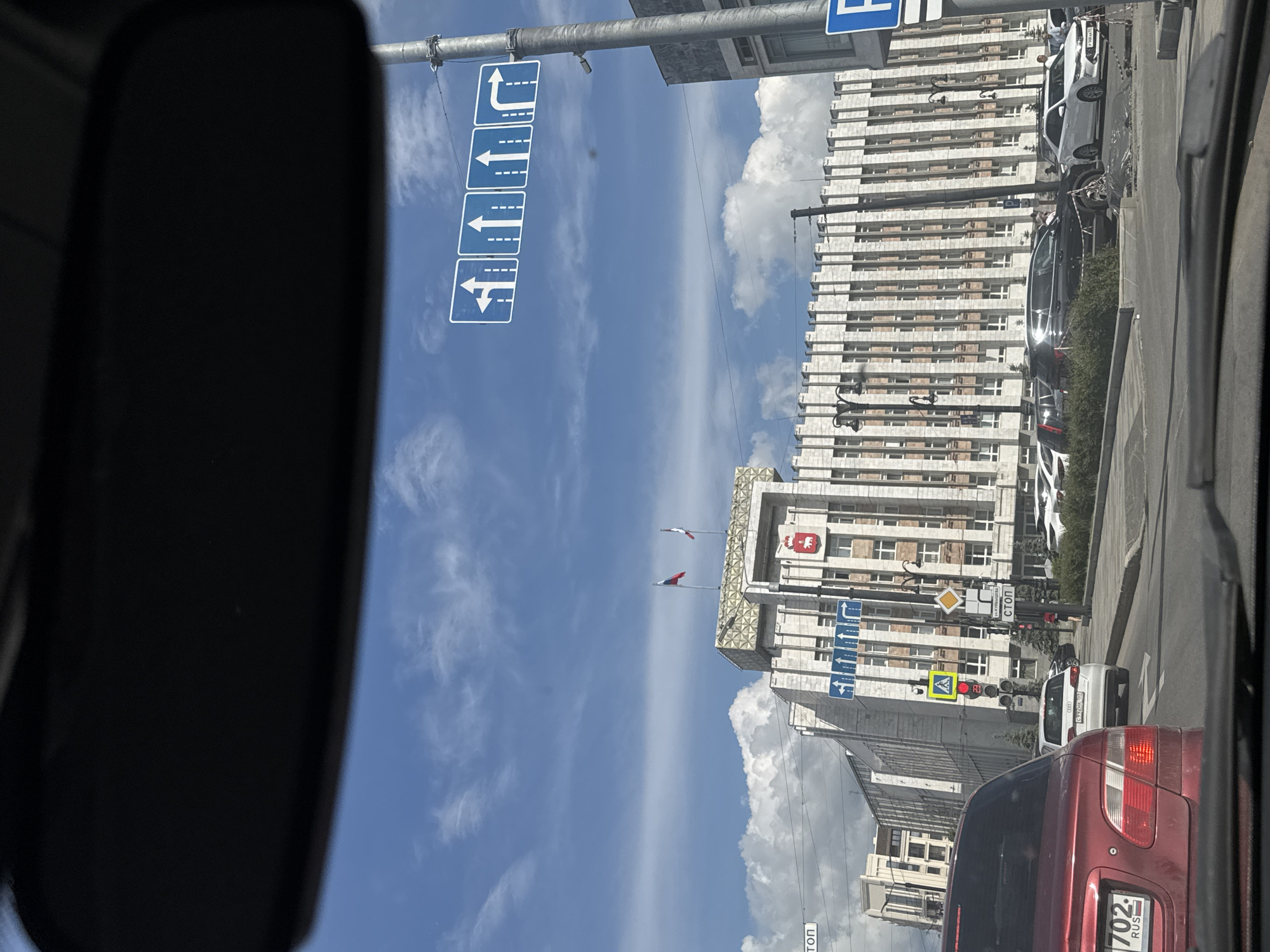 The image, taken from inside a vehicle on a bright, sunny day, depicts an urban street scene in Perm, Russia. A portion of the car's dark rearview mirror is visible on the left side of the frame.
The street is lined with large, multi-story buildings. On the right, a prominent light-colored building with numerous rectangular windows dominates the view. Atop this building, two flags are flying: the Russian national flag (white, blue, red) and a red and white flag, likely representing Perm Krai. Below the flags, a coat of arms, specific to Perm Krai (depicting a red bear carrying a bible with a cross, topped with a crown), is visible on the building's facade, suggesting an official or administrative structure.
Several blue rectangular road signs with white arrows hang overhead, indicating lane directions for drivers: straight ahead, a left turn, a right turn, and a combined straight or left turn. Other road signs, including a yellow diamond-shaped sign for "ГЛАВНАЯ ДОРОГА" (MAIN ROAD) and a red octagonal "СТОП" (STOP) sign, are also present, alongside traffic lights. The sky is a clear blue with scattered white clouds, indicating fair weather.
The foreground features the rear of a red car, displaying a Russian license plate with "702 RUS," confirming the country. Other cars are visible along the street, parked or waiting in traffic. The scene captures a typical urban environment, bustling with road infrastructure and architectural elements characteristic of a Russian city.