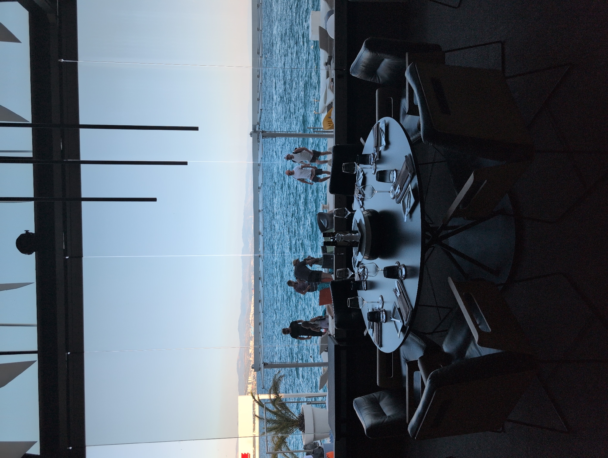 A restaurant dining area features a circular table set for dinner with wine glasses, cutlery, plates, and napkins. Four dark leather chairs surround the table, with two visible in the foreground. The restaurant's large windows offer a view of the sea and a distant coastline, possibly Grad Opatija, Croatia. Outside, several people are visible near the water. A palm tree in a large white planter stands to the left. The sky is clear and blue, indicating daytime.