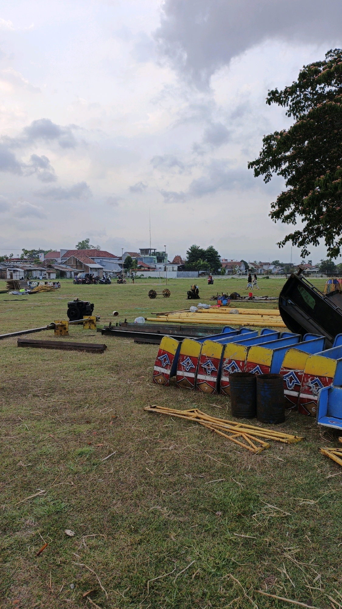In Boyolangu, Indonesia, an outdoor scene unfolds on a grassy field under an overcast sky. Several pieces of equipment and disassembled structures are laid out on the ground, suggesting preparation or disassembly of an event or attraction. In the foreground, several colorful, boat-like seating modules, painted in blue, yellow, and red with decorative patterns, are stacked or arranged. Adjacent to these are dark, cylindrical barrels, possibly for support or ballast, and yellow metal framing structures. Further back, long yellow cylindrical pipes and metal beams are visible, along with what appears to be a disassembled engine or generator. In the middle ground, a few individuals are present, some standing near what could be vehicles or smaller pieces of machinery. The background shows a residential area with houses, some with visible roofs and structures, and a telecommunications tower in the distance. A large tree with dark foliage and some pink blossoms is visible in the upper right corner, partially obscuring the sky. The overall impression is one of an outdoor setup in a semi-rural or town fringe environment.