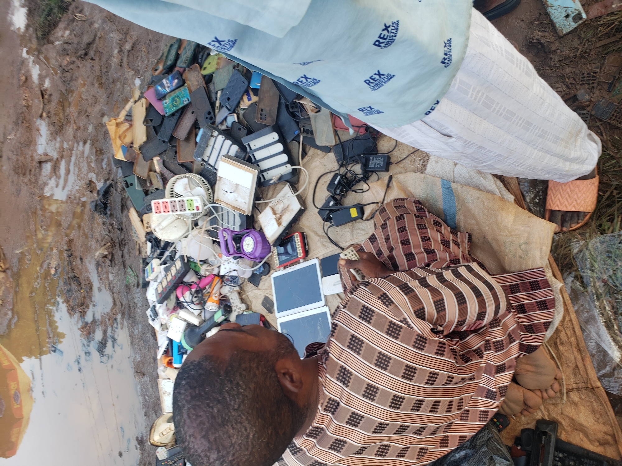 In Kakuri Gwari, Nigeria, an outdoor scene depicts a man, viewed from behind and slightly to the side, examining various electronic devices. He wears a brown and white striped garment and is seated on the ground amidst a collection of discarded electronics. These include multiple mobile phone cases in various colors, chargers, power strips, tablets, and what appear to be old mobile phones. In the background, a muddy, waterlogged area suggests a recent rainfall or a poorly drained environment. Another person, partially visible on the right side of the frame, wears a light blue garment with a repeating "REX CHRISTUS" logo. The overall scene suggests an informal marketplace or a recycling area for electronic waste.