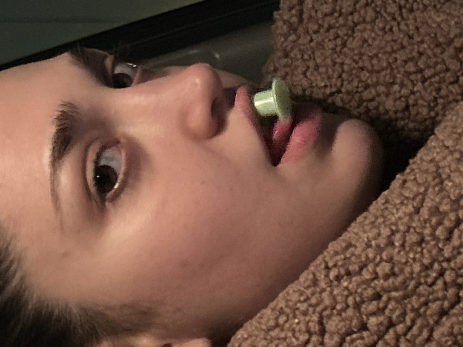 A close-up, angled shot features a young woman's face with a green object held between her lips. Her eyes are wide, conveying a sense of alertness. The skin appears smooth and fair-toned, contrasted by dark eyebrows and hair. A soft, brown, textured fabric is visible near her face, possibly a blanket or jacket. The background is dark, drawing attention to the subject's face.