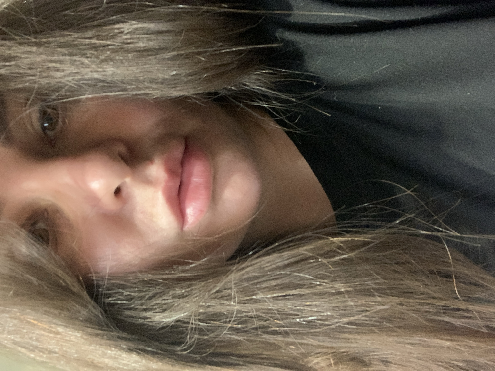 This is a close-up, low-angle, over-the-shoulder shot of a person's face, partially obscured by their long, light brown hair. The person is looking towards the camera with their head tilted. Their eyes are visible, as are their nose and lips, which are slightly parted. The skin appears smooth and fair, with some subtle shine, possibly from makeup or natural oils. The lighting is soft, casting faint shadows that highlight the contours of the face.
The background is dark and out of focus, suggesting an indoor setting with low ambient light. The overall impression is intimate and relaxed, perhaps indicating a candid moment captured casually. The image is somewhat grainy, which is common for photographs taken in low light conditions. No text is visible in the image. The time of day and weather are indeterminate due to the lack of external cues. The emotion conveyed is neutral to slightly contemplative, with the direct gaze at the camera being the primary indicator.