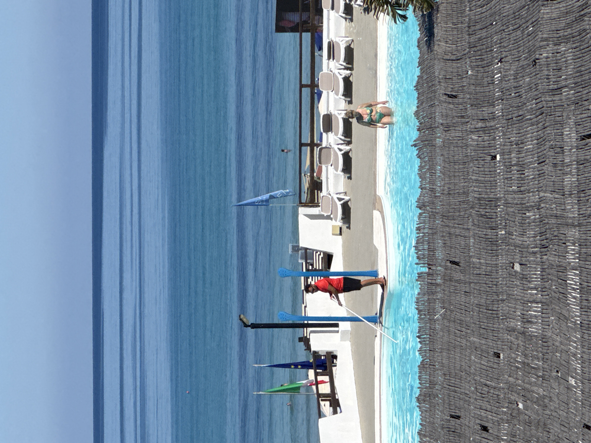 A lifeguard in a red shirt and black shorts uses a pool skimmer to clean a bright blue swimming pool. A woman in a green and yellow bikini stands in the pool. In the background, the Mediterranean Sea meets a clear blue sky. Several lounge chairs are visible near the pool, and flags representing Italy and the European Union are planted in the ground. The scene takes place at a seaside resort in Isola delle Femmine, Italy.
