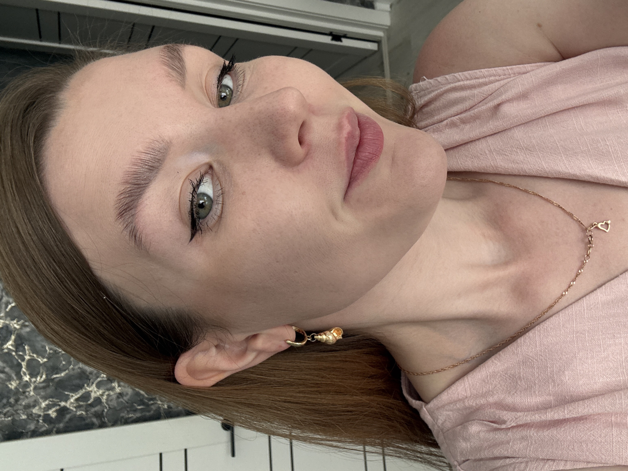 A medium-shot of a fair-skinned person with light brown hair, facing the camera at an angle. They have light green eyes, noticeable freckles, and are wearing dark eyeliner. Their eyebrows are full. They are wearing a light pink, off-the-shoulder top. A gold necklace with a small heart pendant is visible, as is a gold earring shaped like a seashell. The background includes a white and gray patterned surface and white vertical bars. The location is unknown and could not be determined to be within the city of Izhevsk, Russia.