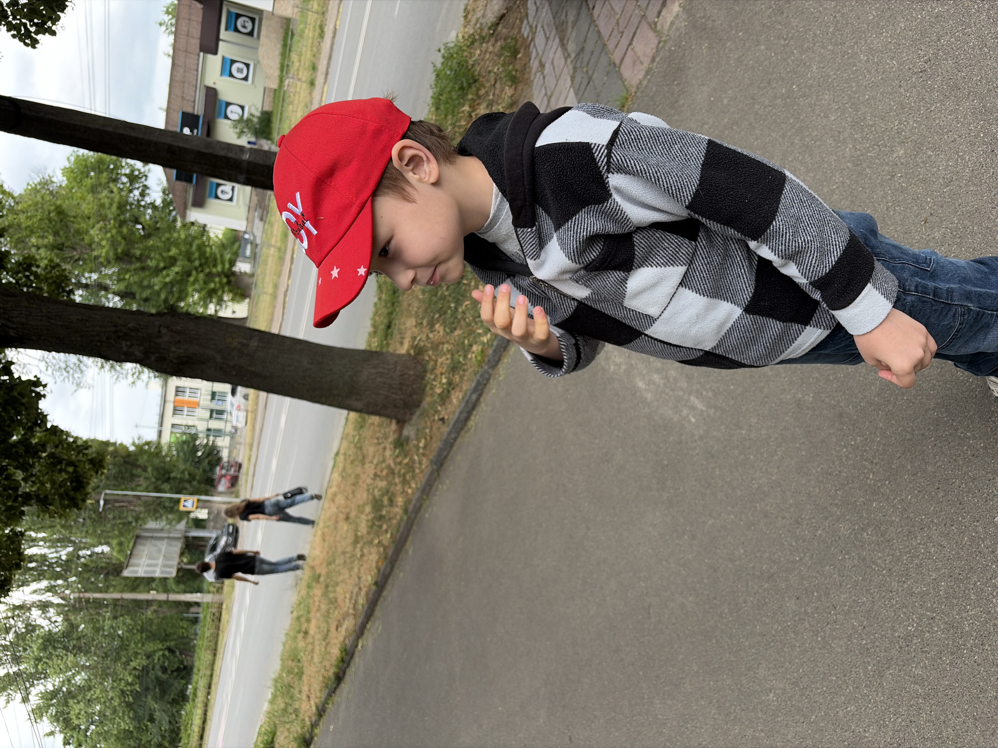 A young boy wearing a red baseball cap with white stars and a black and white checkered hooded jacket stands on a paved sidewalk. He is facing to his right, looking away from the camera with his left hand raised to his chin. Behind him, across a road with two lanes of traffic, are buildings and trees. A man and a woman, dressed in casual clothing, are walking away from the camera on the sidewalk across the street. The location is identified as Chernihiv, Ukraine.