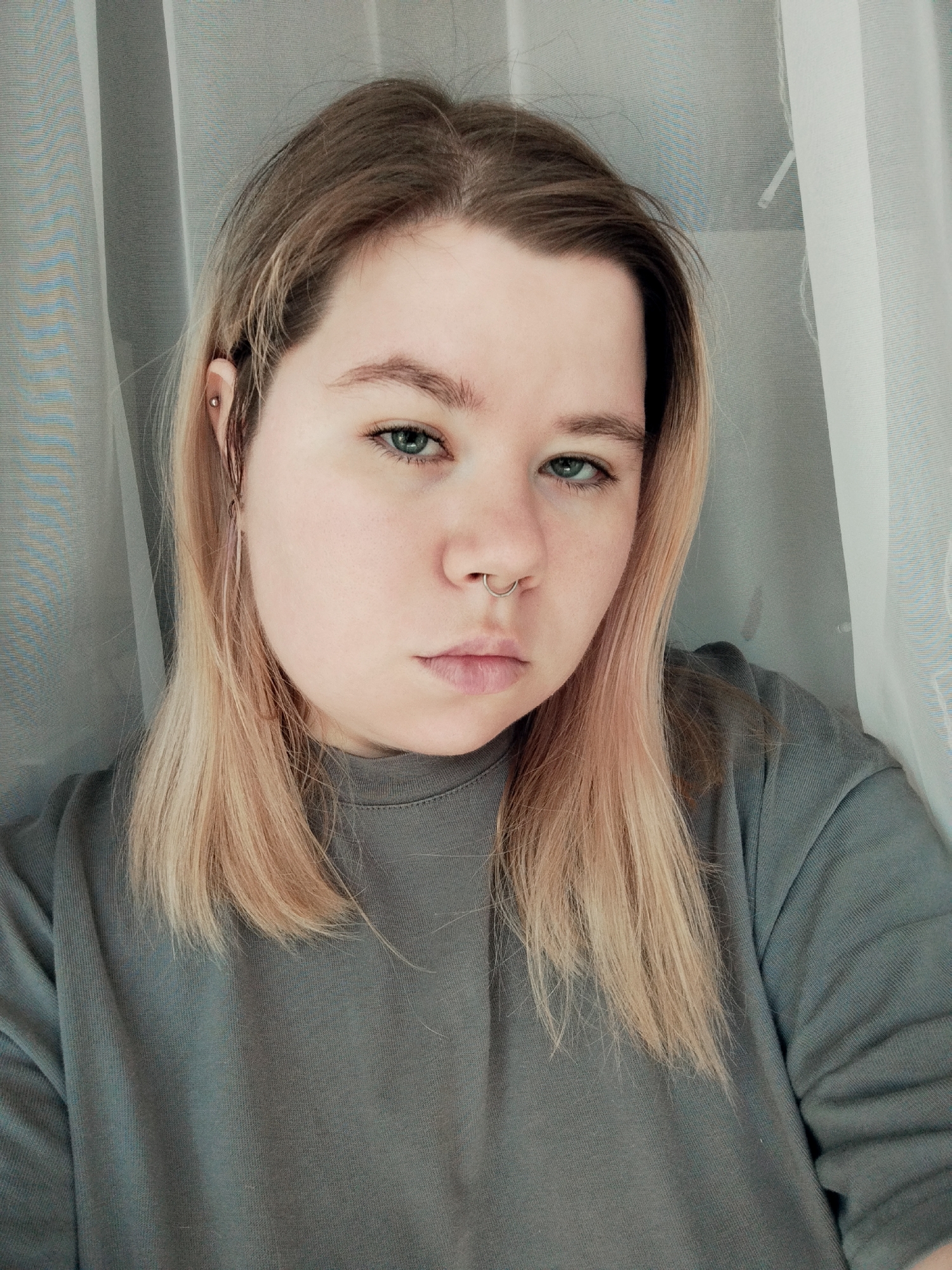A medium-shot portrait shows a fair-skinned individual wearing a gray long-sleeved shirt, with long light brown hair, and a septum piercing. The person is positioned in front of sheer white curtains. A small stud is visible in the upper ear. The location is Unknown, Unknown.