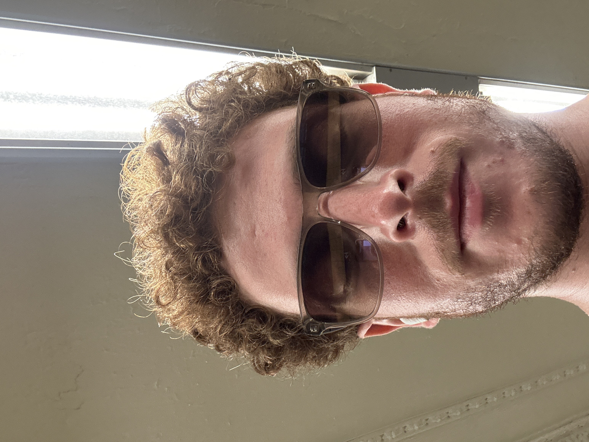 A close-up, tilted image shows a person with curly brown hair, sunglasses, and a beard. The individual is under fluorescent lighting. The location cannot be determined from the image.