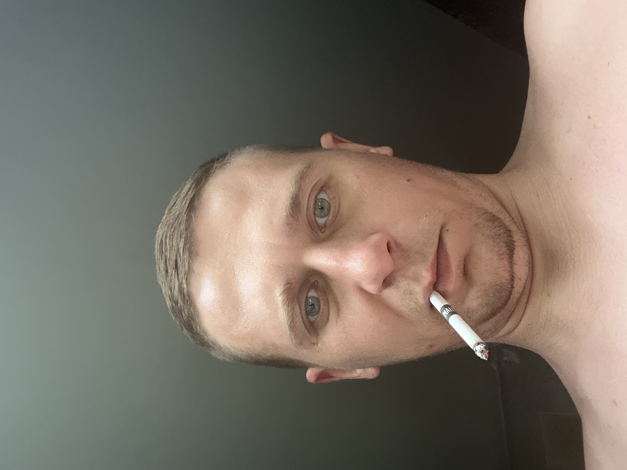 A bare-chested male individual with short, light-colored hair and light-colored eyes is depicted in a close-up, low-angle photograph taken in the city of Minsk, Belarus. He has light stubble on his chin and upper lip, and a lit cigarette, with visible "MAL" branding on the filter, is held between his lips. The tip of the cigarette shows glowing embers. The subject looks directly into the camera with a neutral expression. The background consists of a plain, uniformly dark grey-green surface, indicating an indoor setting with soft, indirect lighting from above.