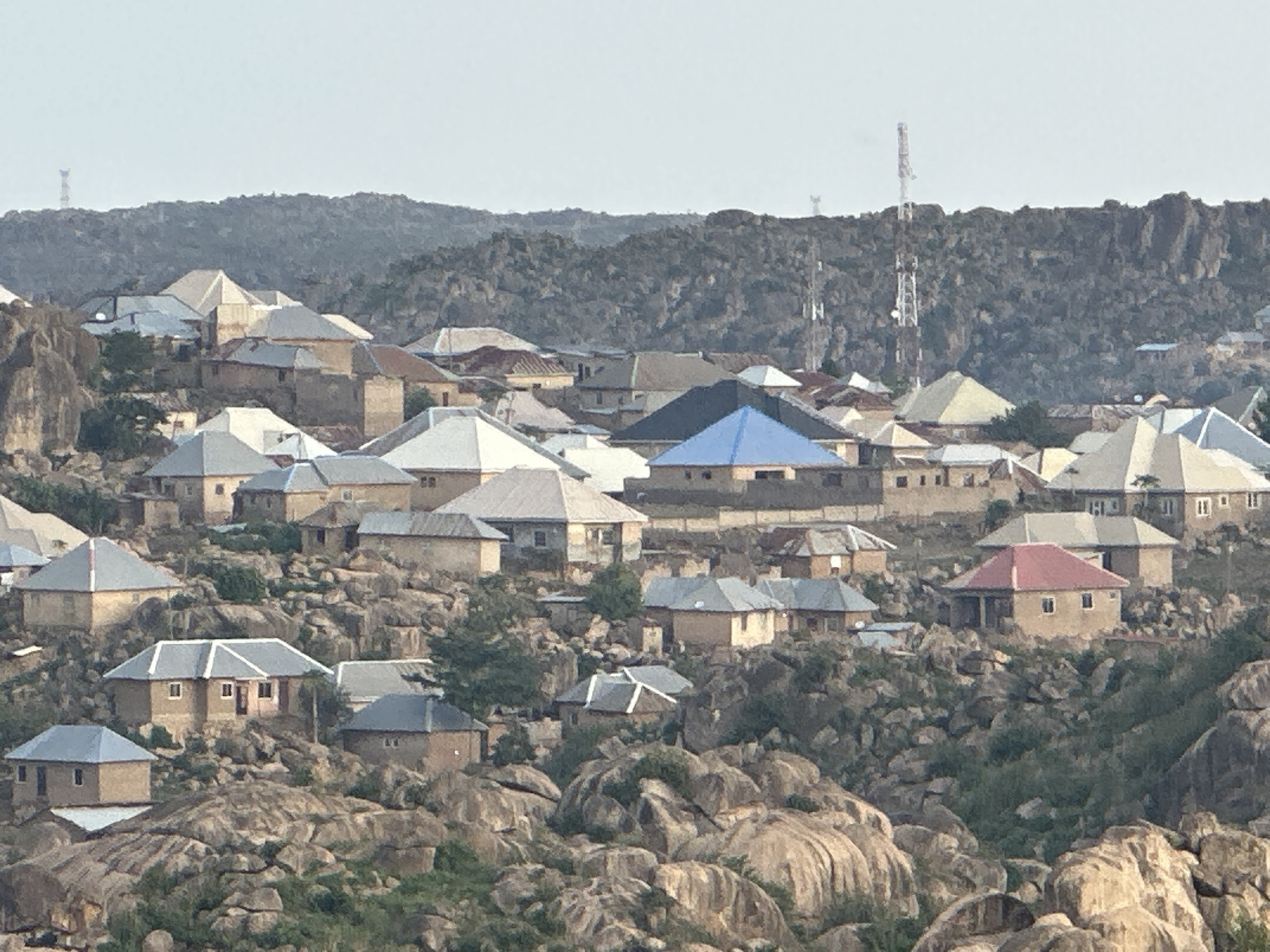 The image shows a densely populated residential area in Jos, Nigeria, characterized by buildings with diverse roof colors (gray, blue, red, white) constructed on a rocky, uneven terrain. The houses are generally single-story and simple in design. The landscape is dominated by large boulders and sparse vegetation. In the background, a ridge of rocky hills is visible, dotted with communication towers. The sky is overcast. There are no people visible in the scene. The overall impression is of a community built in harmony with a rugged, natural environment.