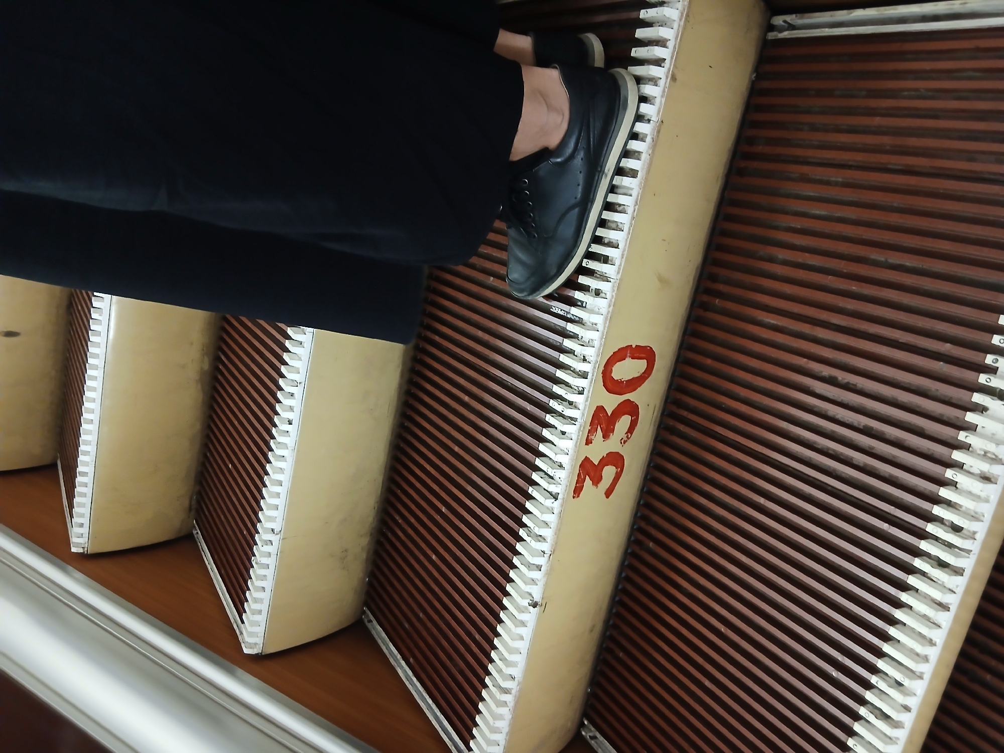 The image shows a close-up view of an escalator. The steps are made of dark brown slats with white comb-like edges. A person, wearing black pants and black shoes, is standing on one of the steps. The number "330" is painted in red on the side of one of the escalator steps. The escalator appears to be indoors, with a wooden paneling on the side and a silver handrail visible in the upper left corner. The perspective is from a low angle, looking up the escalator.