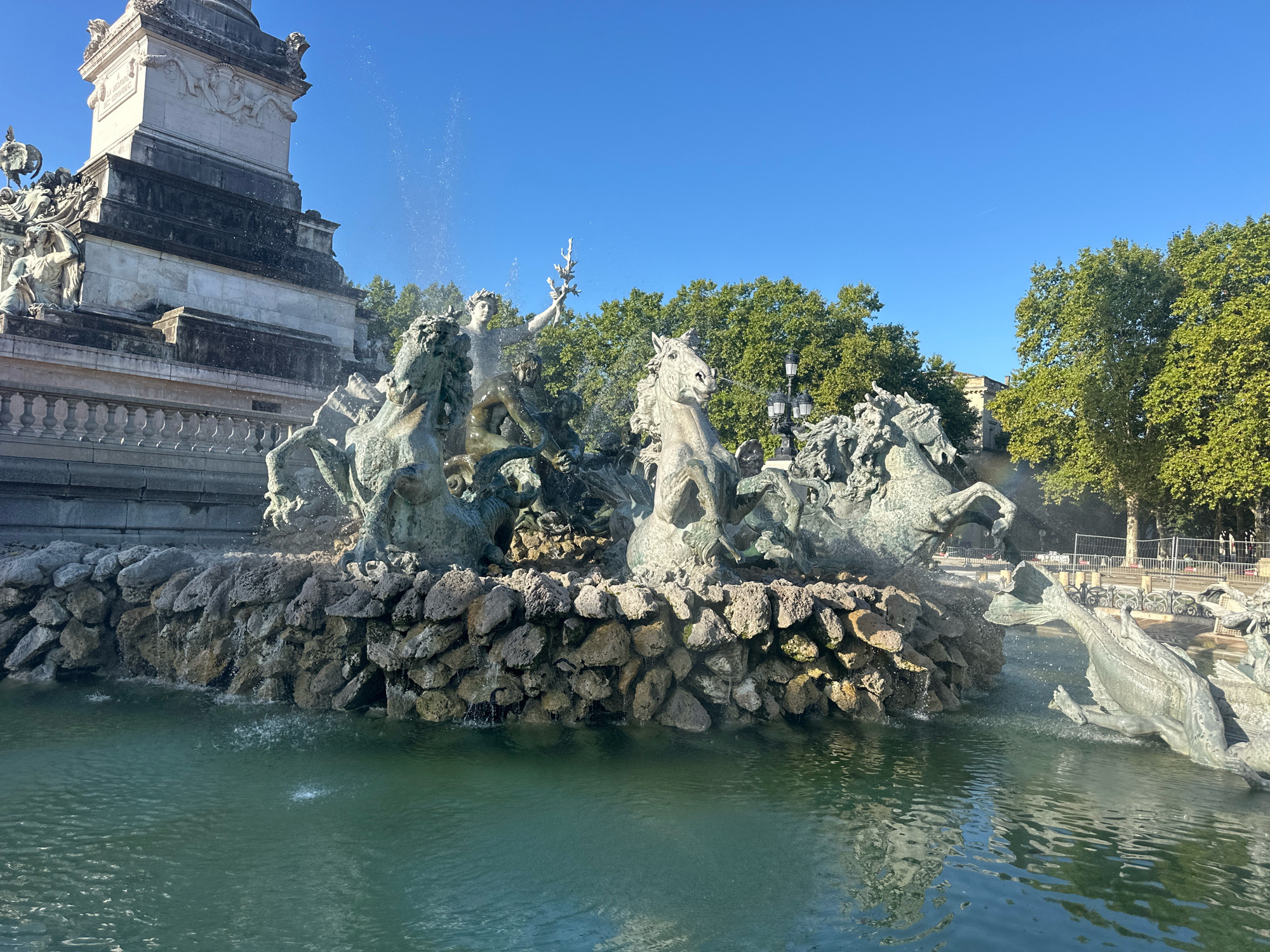 The image shows the monumental fountain of the Place des Quinconces in Bordeaux, France, on a clear, sunny day. The fountain features several bronze sculptures of horses and mythological figures, with water spraying from various points. In the background, a large stone monument stands to the left, and lush green trees line the perimeter of the square. The water in the fountain is a deep green color, reflecting the sky and surrounding structures. There are no people clearly visible in the frame.