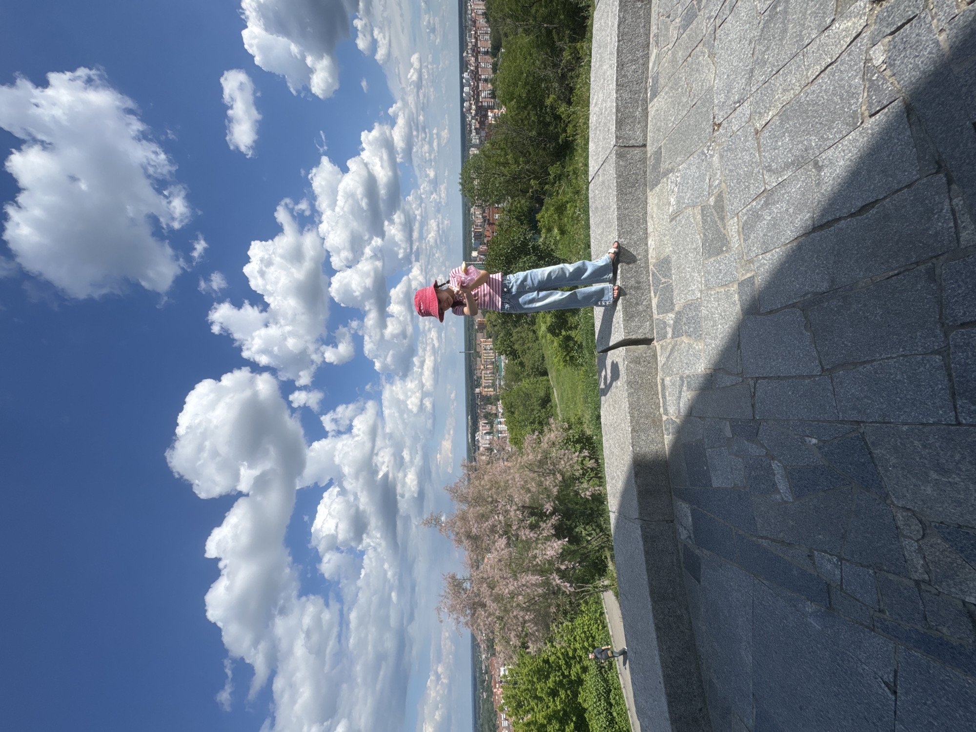 The image displays a sunny daytime scene from an elevated perspective, overlooking a distant city and green landscape. In the mid-ground, a person, likely a young woman or girl, stands on a grey stone-paved platform. She is wearing a red and white striped shirt, blue jeans, and a red bucket hat, and appears to be looking down at something in her hands.
The foreground and much of the mid-ground are covered by this stone paving, which consists of large, irregular slabs. Beyond the platform and the person, a lush green area unfolds, featuring numerous trees and bushes. A prominent tree with light pink or purple blooms stands out against the green foliage. In the background, a sprawling cityscape is visible under a clear blue sky, dotted with numerous white, fluffy cumulus clouds. Shadows are cast across parts of the stone ground, indicating bright sunlight. The scene suggests a pleasant day at a scenic overlook or park. No text is visible in the image.