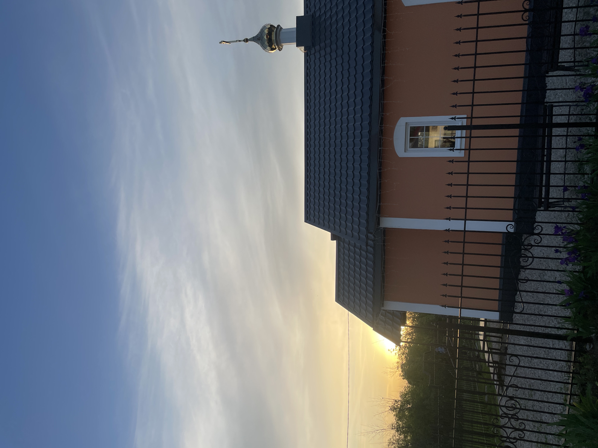 A photograph depicts a portion of a building, possibly a church, in Saratov, Russia. The building has an orange facade, a dark gray roof made of tiles, and a small window with white trim visible behind a black metal fence. A thin steeple extends from the top of the roof. The sky is pale with yellow hues. The fence runs along the building's perimeter, and vegetation is visible beyond it. The sun is setting, with the light reflecting off the sky and the building.