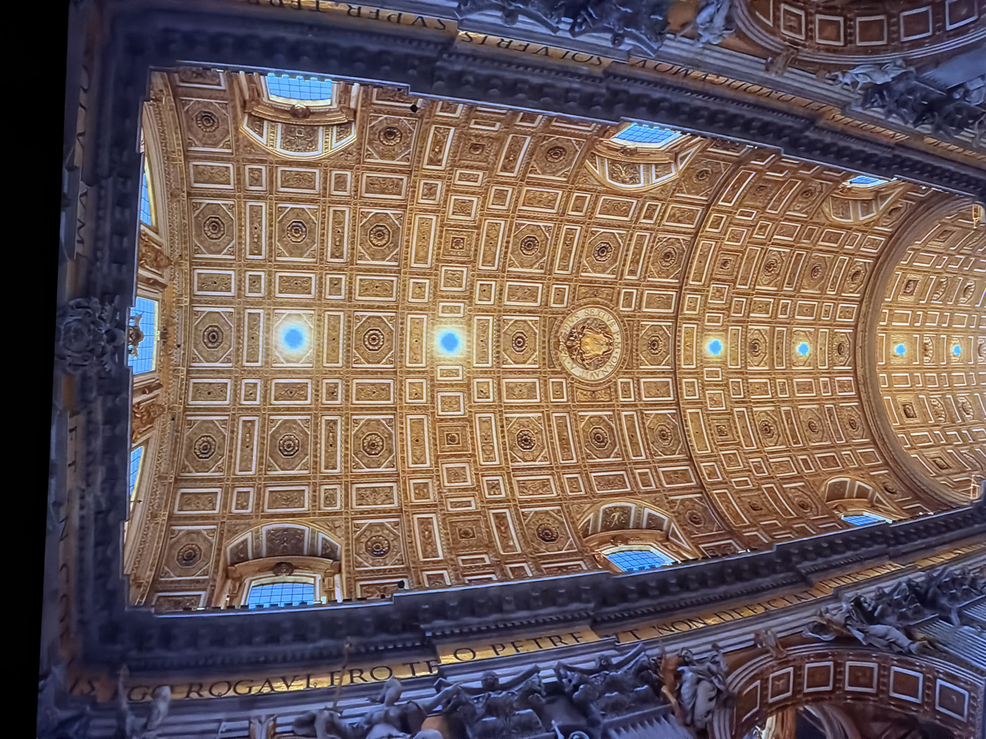 The image shows a highly ornate, gold-colored ceiling with a repeating geometric pattern of squares and octagons, likely in a religious or historical building. There are multiple small, bright blue illuminated elements scattered across the ceiling. The sides of the image reveal architectural details, including dark, textured borders and decorative statues or figures. The overall impression is of grandeur and intricate craftsmanship. The image does not contain any elements that would place it in the city of Chirchik, Uzbekistan.