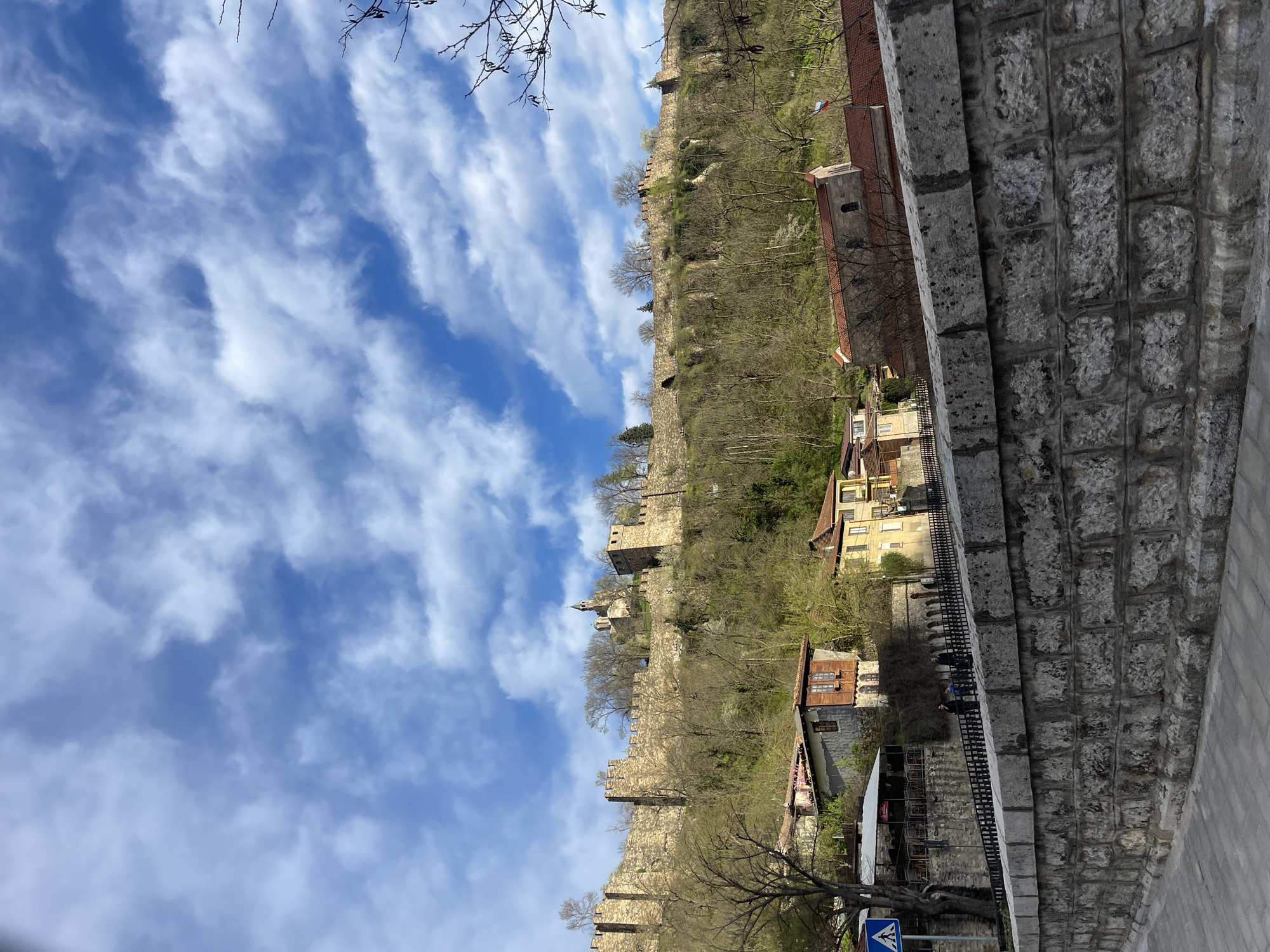The image depicts a view in Veliko Tarnovo, Bulgaria, featuring a stone wall and fortifications along a hill. In the background, residential buildings nestle amongst trees. Above, a blue sky is partially obscured by wispy clouds. A stone structure dominates the right foreground. A crosswalk sign is visible.