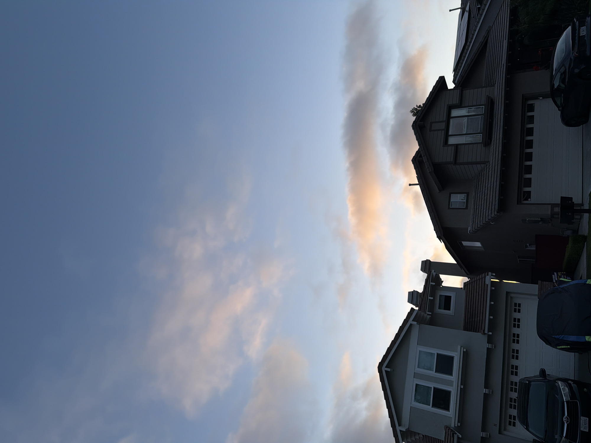 The image shows a residential street in Aliso Viejo, California, USA at dusk. Two modern houses with garages are visible, with a clear sky and scattered clouds above. A dark-colored SUV is parked in front of the house on the left, and another dark-colored vehicle, possibly a sports car, is parked in front of the house on the right. One of the garages is covered with a dark tarp. Solar panels are visible on the roof of the house on the right. The overall impression is of a quiet suburban evening.