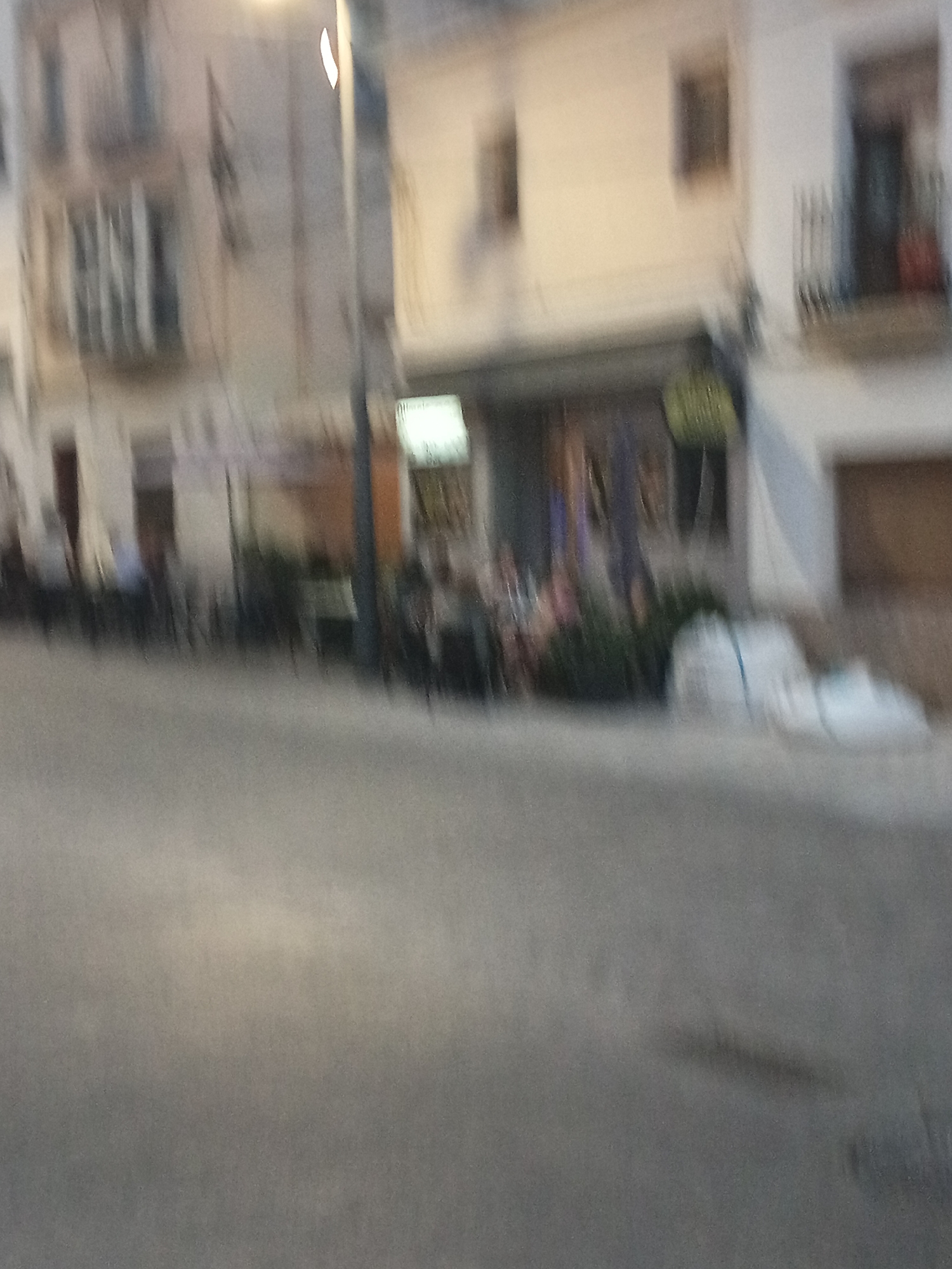 The image depicts a blurred street scene in Tortosa, Spain, likely during dusk or evening. In the background, buildings with multiple windows and balconies line the street. One building appears to have a shop or cafe on the ground floor with tables and chairs set up outside. Several indistinct figures are seated at these tables, suggesting outdoor dining or socializing. Further down the street, more seating areas are visible, some with umbrellas. White plastic bags or coverings are piled near the buildings, potentially for storage or waste. The foreground is dominated by a paved street with a slightly reflective surface. The overall impression is of a quiet urban environment, though the blurriness of the image makes precise identification of details challenging.