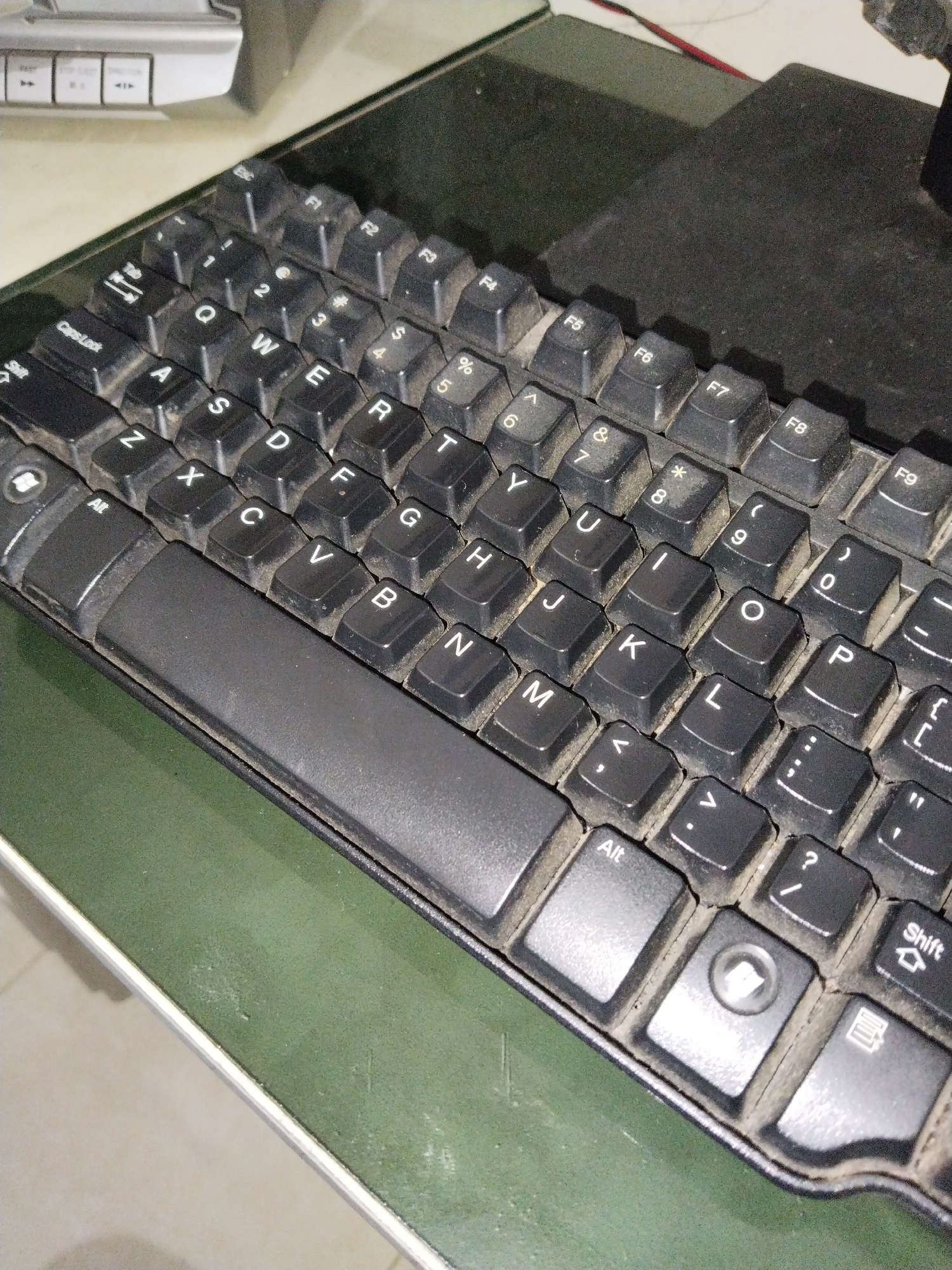 The image shows a close-up, top-down view of a black keyboard covered in dust. The keyboard is placed on a green surface, possibly a desk or table. The keys are visible with their standard QWERTY layout, along with some function keys at the top. Some of the keys are highlighted with their characters. The overall scene suggests a dusty environment, common in some indoor settings. The image does not contain any information about the location being Banyurojo, Indonesia.