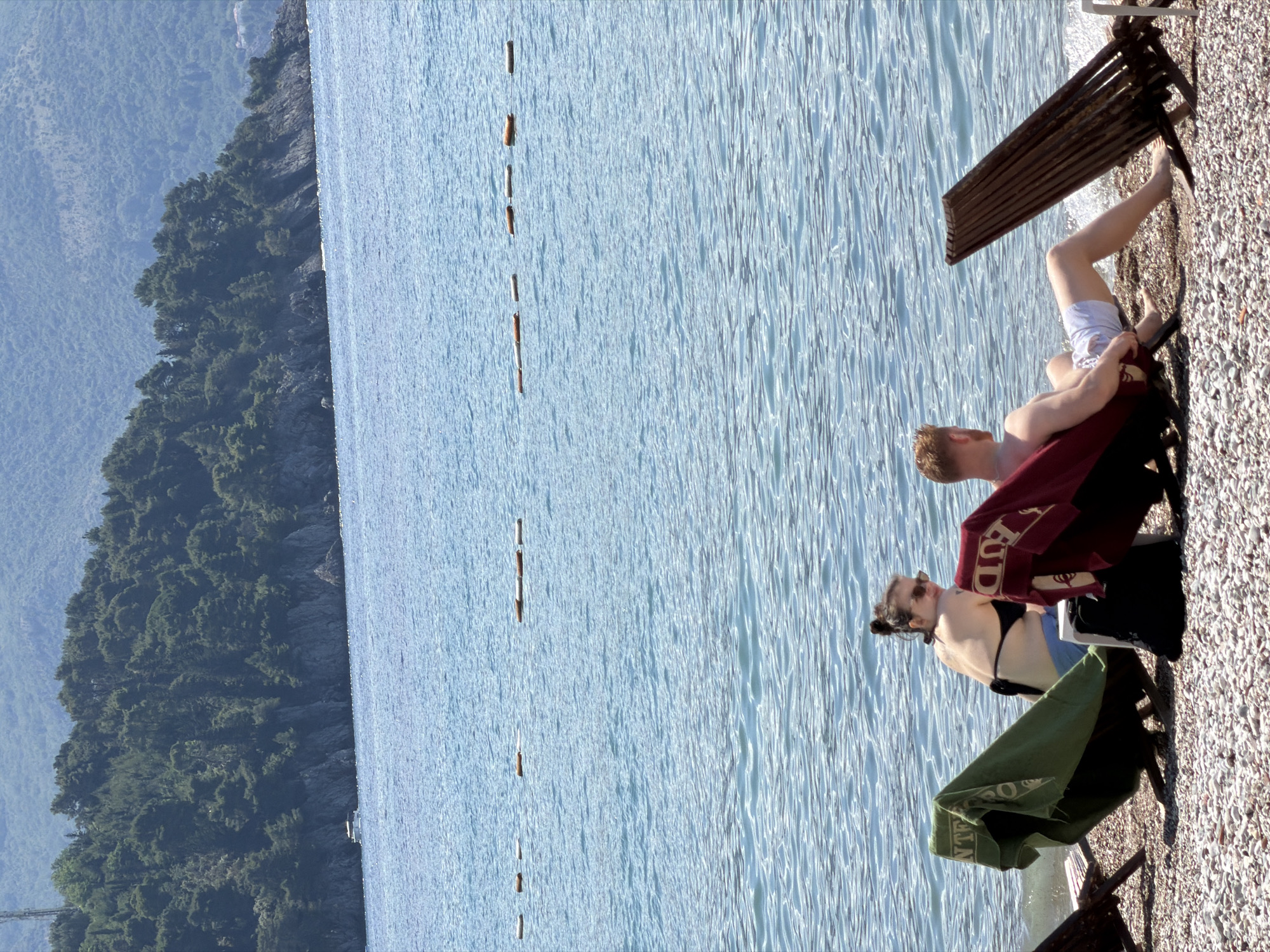 The image captures a sunny beach scene in Budva, Montenegro, featuring two individuals relaxing by the sea. In the foreground, on a pebble beach, a man and a woman are seated on sun loungers. The man, partially visible, wears white shorts and is leaning back, with a red towel bearing the text "BUD" (likely for Budva) draped over his chair. Behind him, a woman wearing a black bikini top and sunglasses is also seated on a lounger, covered by a green towel with the text "NTE" (possibly for Montenegro).
To their left, the clear blue sea stretches out, showing gentle ripples on its surface. A line of small, brown buoys or markers extends into the water, suggesting a designated swimming area. In the background, a rugged coastline covered densely with green trees and foliage rises, transitioning into distant, hazy mountains. The bright sunlight indicates it is daytime with clear weather. The overall impression is one of leisure and relaxation by the Mediterranean coast.