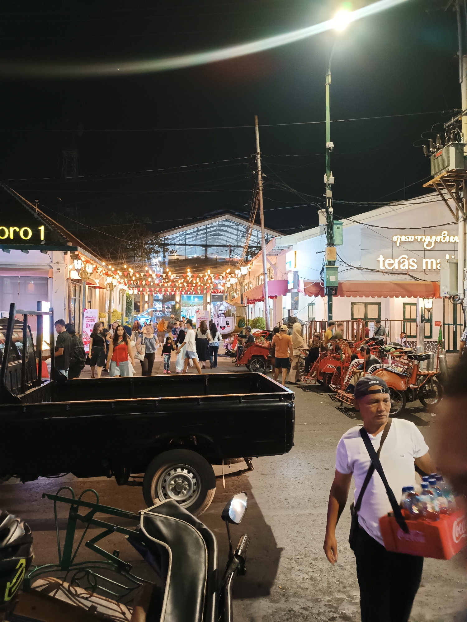 The image depicts a busy street scene at night in Yogyakarta, Indonesia, likely a tourist area. A man in a white t-shirt and black pants walks towards the viewer, carrying a red carrier with bottles of water. In the foreground, a black pickup truck is parked on the left, and a part of a motorcycle is visible on the right. The street is illuminated by string lights and a street lamp.
Several people are walking on the sidewalk, browsing shops, and gathering around red trishaws, also known as "becak," which are a common mode of transport in Indonesia. Buildings line both sides of the street, with some displaying signs like "tiras ma'" and "pro 1." The atmosphere appears lively and crowded, suggesting a marketplace or a night market. The architecture of the buildings suggests a blend of modern and traditional elements. The overall scene is characteristic of a vibrant urban environment in Yogyakarta at night, possibly around Malioboro street, known for its shops, street food, and cultural attractions.