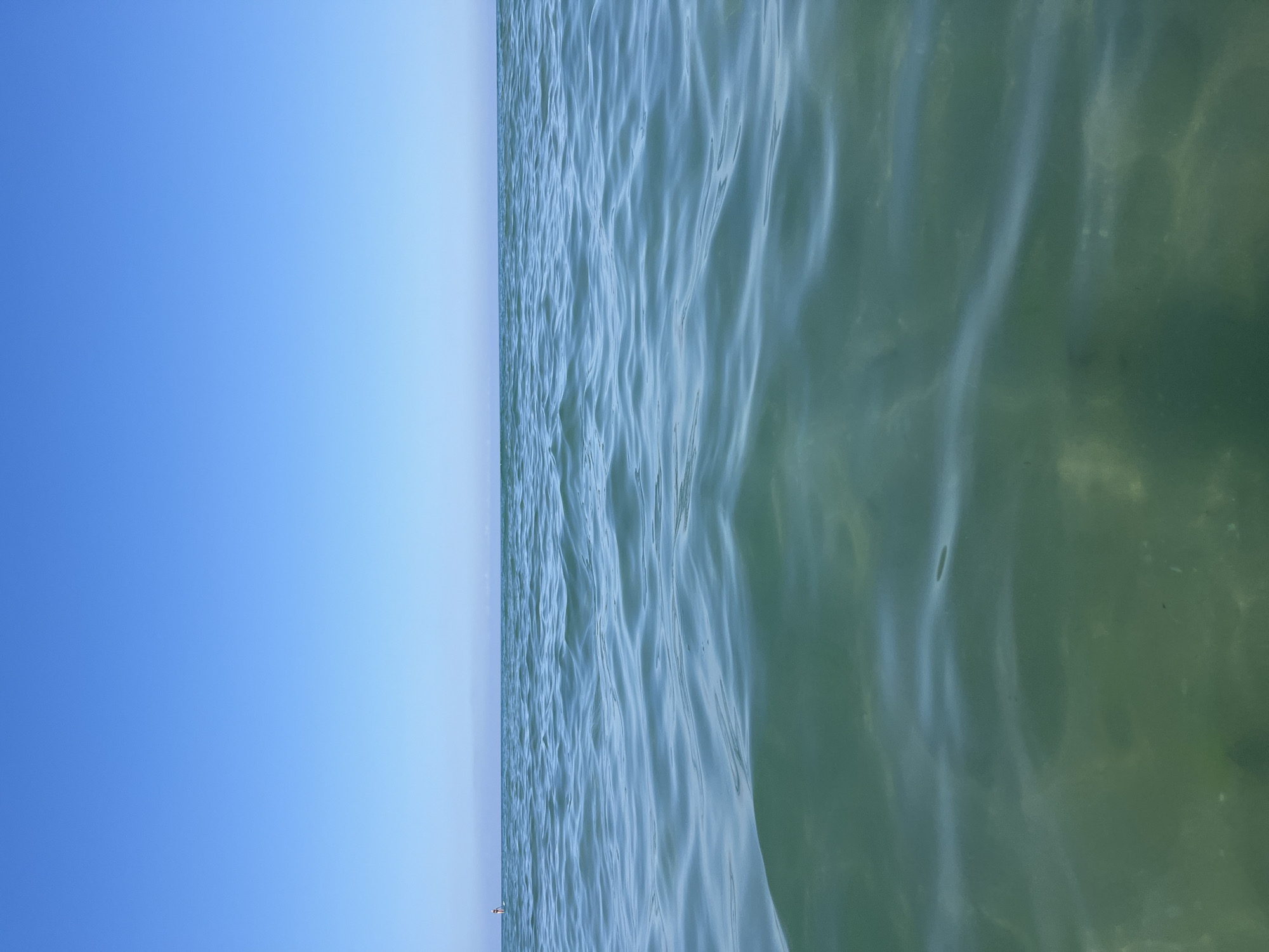 The image depicts a tranquil scene of the sea under a clear blue sky. The horizon line is distinct, separating the vast expanse of water from the sky above. The water is a clear, greenish-blue, with gentle ripples creating subtle patterns on its surface. The sunlight filters through the water, illuminating the sandy or silty bottom in some areas, giving it a translucent appearance. In the distance, on the horizon, a single figure can be seen on what appears to be a paddleboard. The environment suggests a calm, open body of water, likely a coastal area. There is no indication of the city of Emona, Bulgaria, in this image, as the scene is entirely maritime and devoid of urban features.