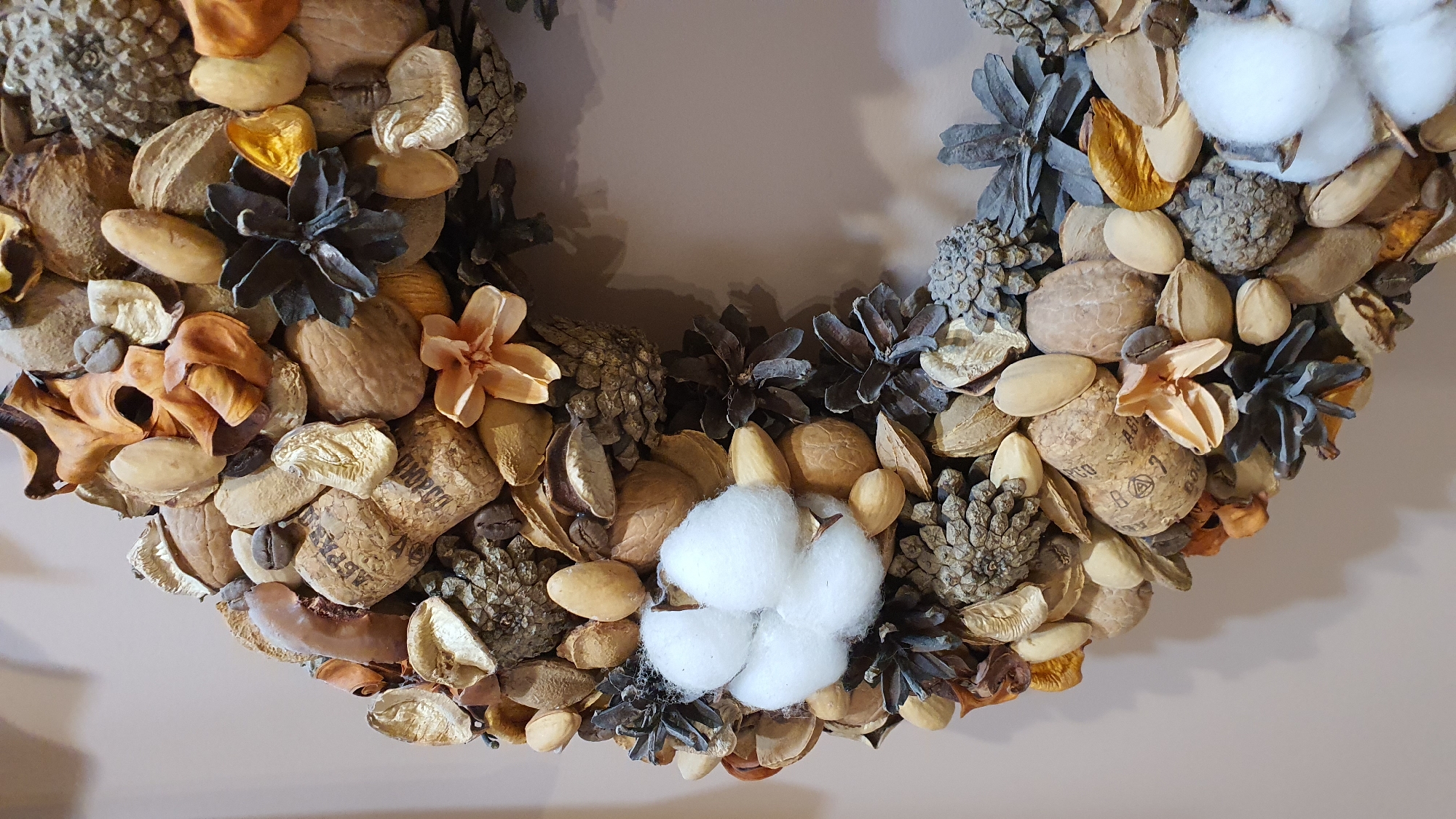 A close-up, slightly angled shot shows a portion of a circular wreath made from natural elements. The wreath is densely packed with various organic materials in shades of brown, beige, and gray. Visible components include nuts (walnuts and almonds), pine cones, dried seed pods, small dried flowers or leaves, and what appear to be coffee beans. A prominent feature is a cluster of fluffy white cotton bolls. One section of the wreath includes a wine cork with visible text. The background is a plain, light beige wall. There are no people or animals present. The image was taken indoors, likely as a decorative item, and there is no discernible connection to the city of Krasnoyarsk, Russia.