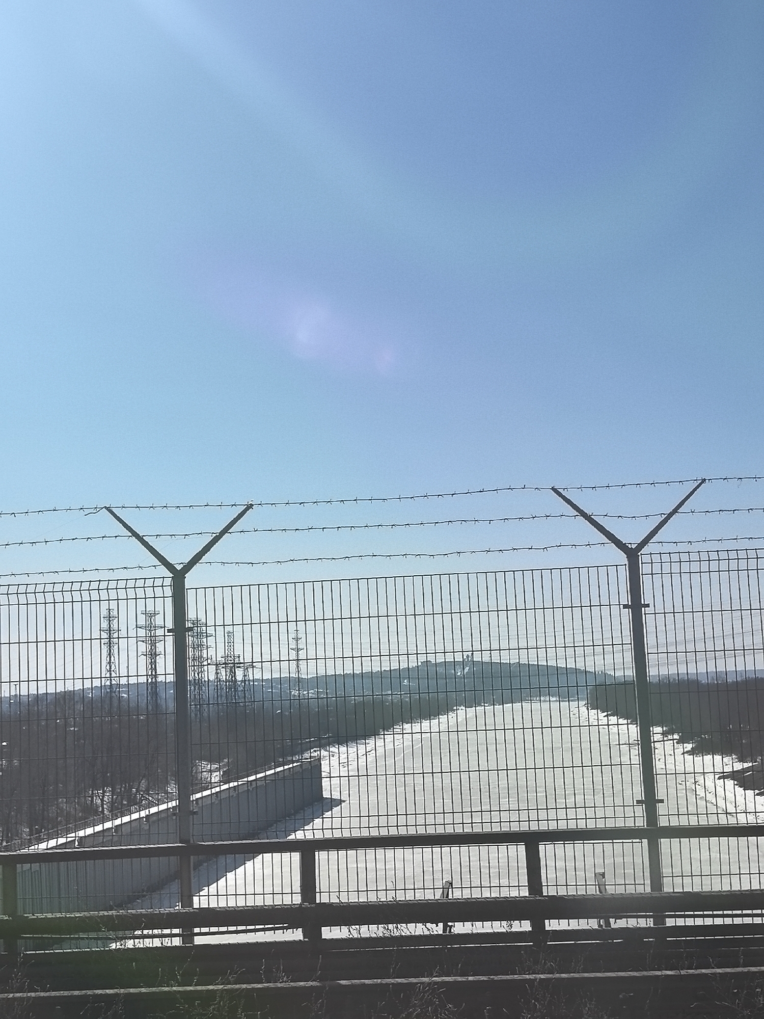 The image shows a view through a chain-link fence with barbed wire on top, likely a security measure, in Chaikovsky, Russia. Beyond the fence, a wide, empty space or runway is visible, bordered by concrete structures. In the distance, power lines and pylons are visible against a clear, bright blue sky. The ground around the fence is sparse with some vegetation.