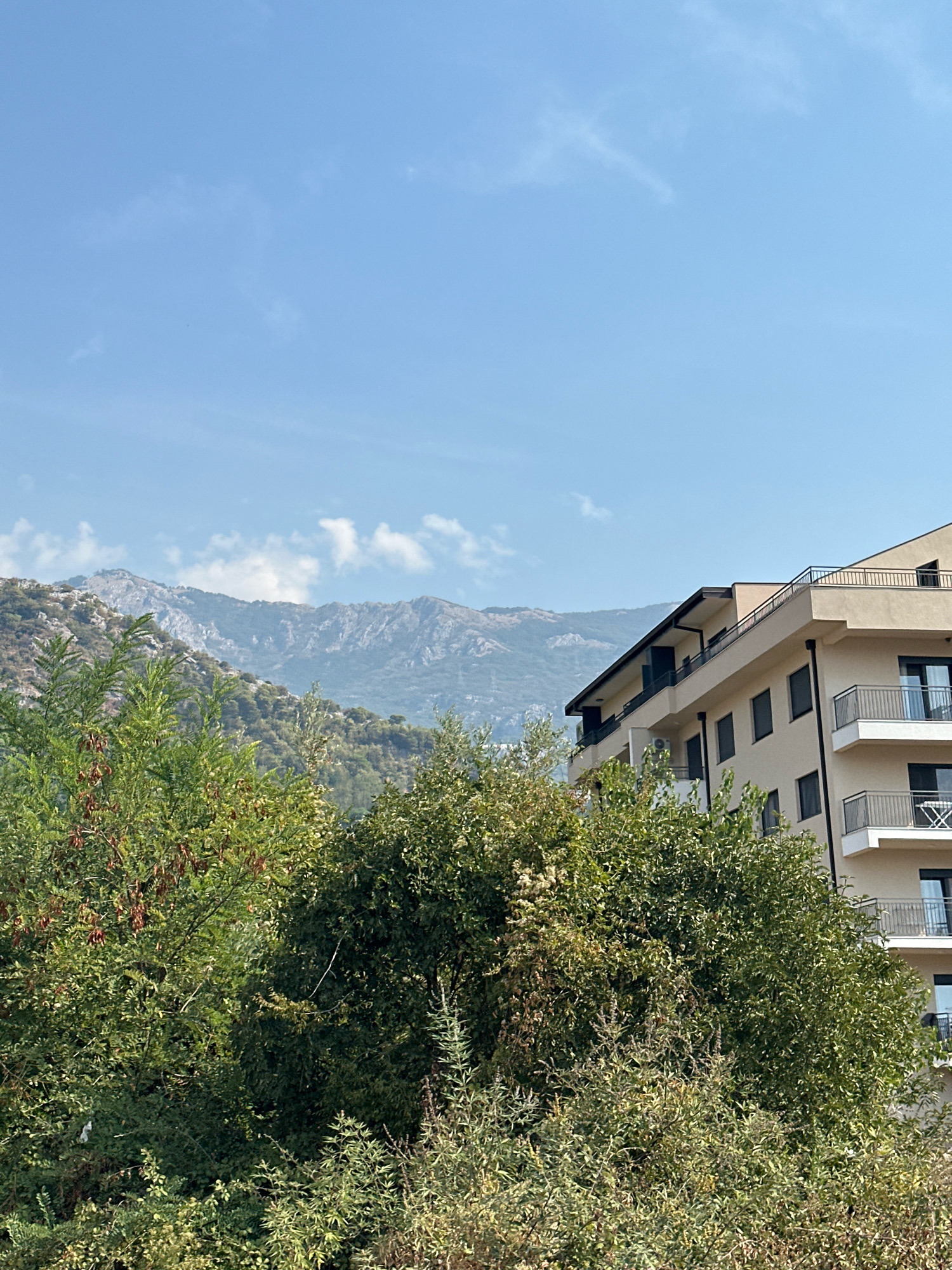The image depicts a view of a modern apartment building nestled against a backdrop of rugged, mountainous terrain under a clear blue sky with scattered white clouds. The building is a light beige color with dark-framed windows and multiple balconies with metal railings. Lush green trees and dense shrubbery partially obscure the lower floors of the building, creating a sense of natural integration with the urban structure. In the distance, the mountains rise, characterized by rocky slopes covered in sparse vegetation, suggesting a dry or arid climate. The overall scene conveys a peaceful, scenic setting, likely in a coastal or semi-mountainous region. The location is identified as Budva, Montenegro.