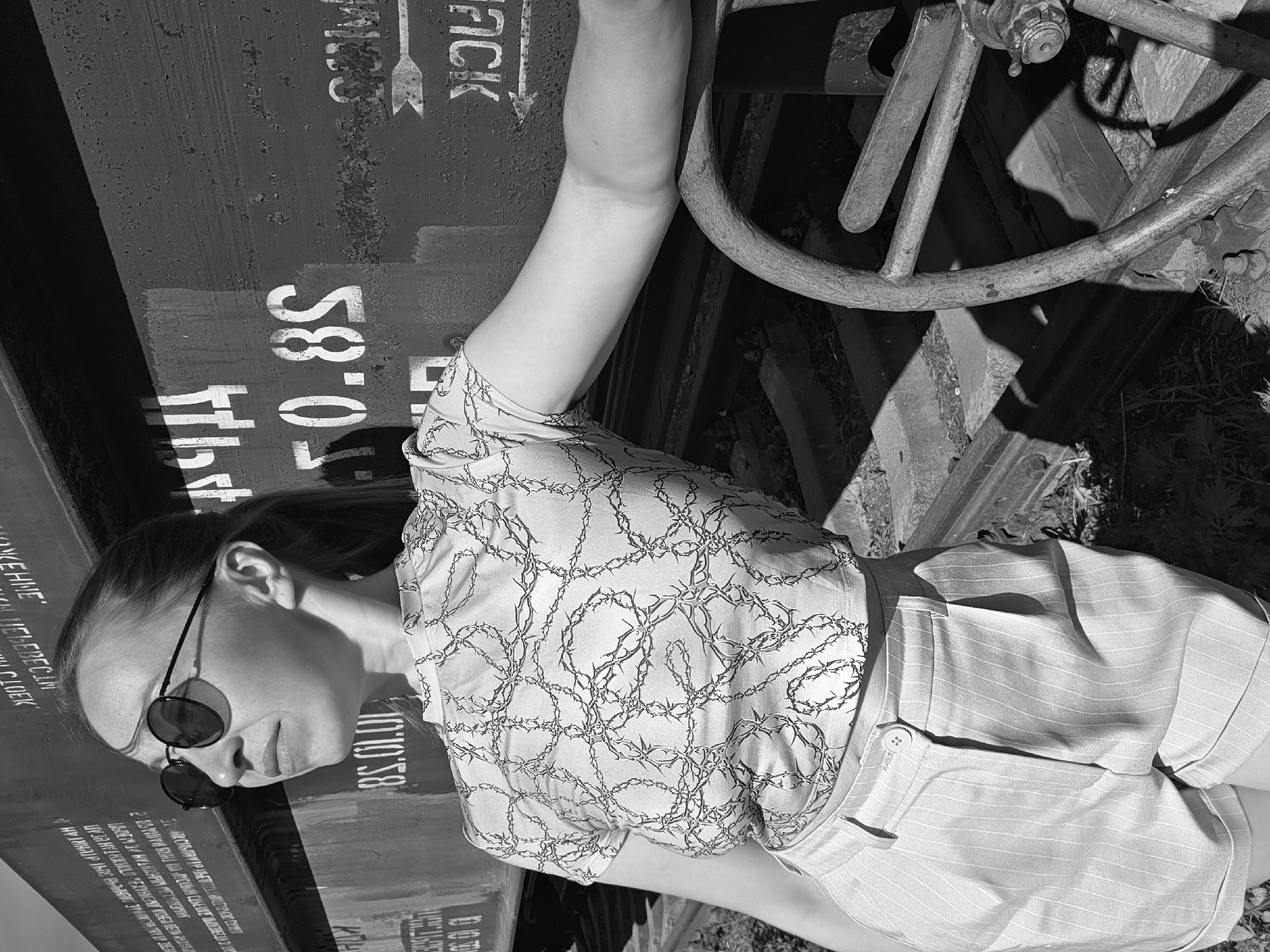 The image is a black and white medium shot of a young woman with light skin and dark sunglasses, wearing a patterned top and shorts, leaning against a weathered train car in what appears to be an industrial or rail yard setting. The train car has white stenciled markings, including Russian text and numerical designations, indicating that it is likely located in Russia. Based on information from the OCR text, the location is likely near or in the city of Chaikovsky. The woman's arm rests on a large metal wheel, possibly part of the train car mechanism. There are traces of vegetation on the ground, suggesting the area is somewhat unkempt or abandoned. The lighting is strong, with distinct shadows adding contrast. The overall impression is a juxtaposition of the woman’s casual attire with the rough, industrial backdrop.