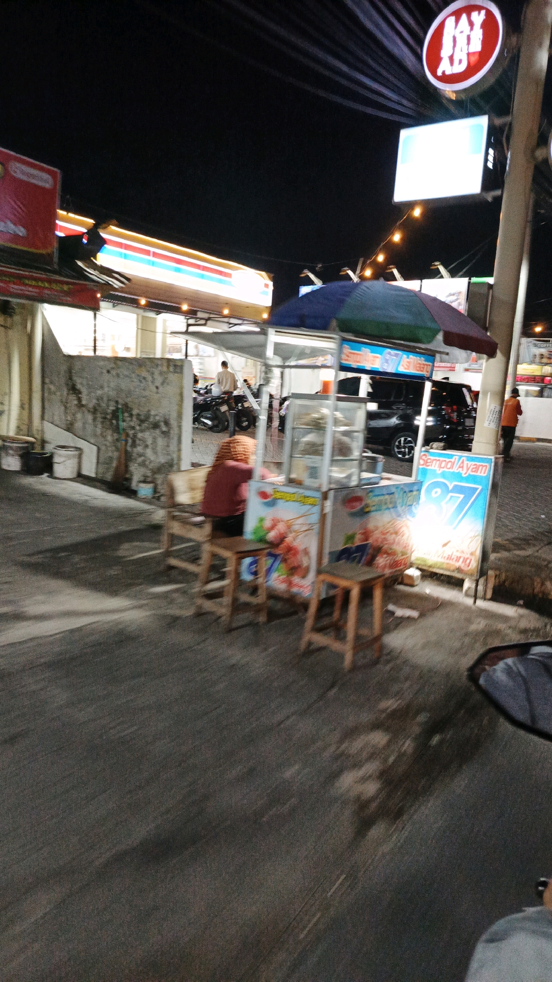 This is a nighttime street scene in Pabelan, Indonesia, featuring a food stall and a convenience store. A woman wearing a red jacket and headscarf sits on a wooden stool in front of a food cart that displays images of skewered food and the text "Sempal Ayam" and "87". The cart is partially covered by a blue and green umbrella. Behind the food stall, a convenience store with bright lighting is visible, with its signage displaying "JAY AB" in a red circle. Several motorcycles are parked near the convenience store. A black SUV is parked on the street in front of the store. The street itself is paved and appears wet in places, with faint road markings. The overall atmosphere is that of a commercial area at night, with activity centered around the food stall and the convenience store.
